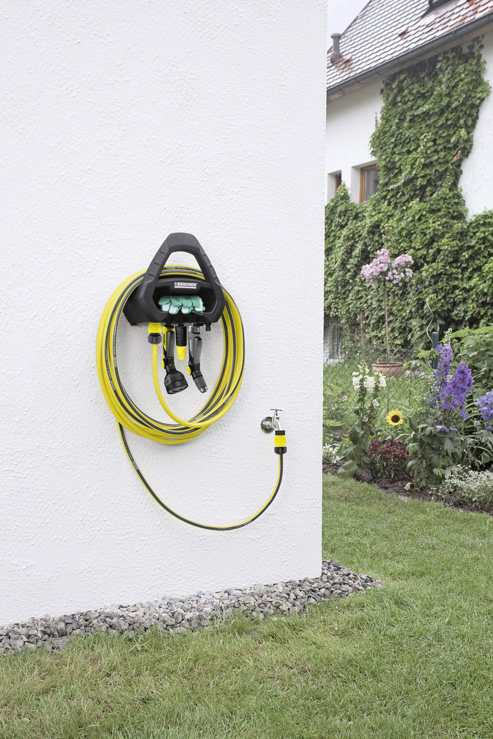 Ein gelber Gartenschlauch ist an einem Halter an einer weißen Hauswand aufgerollt. Im Hintergrund ist ein blühender Garten sichtbar.