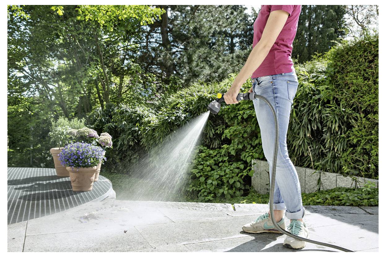 Eine Person in Jeans und einem pinken Hemd bewässert Pflanzen mit einem Gartenschlauch in einem Garten, umgeben von grünem Blattwerk und Topfblumen.