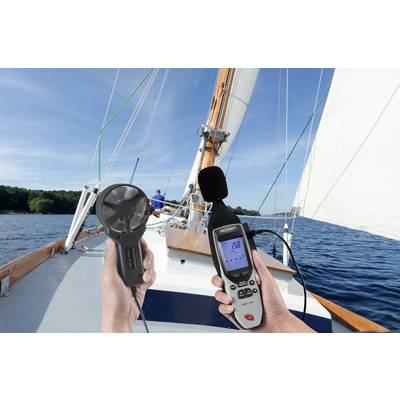 Eine Person auf einem Segelboot misst mit einem Anemometer die Windgeschwindigkeit. Das Boot segelt bei klarem Himmel auf einem See.