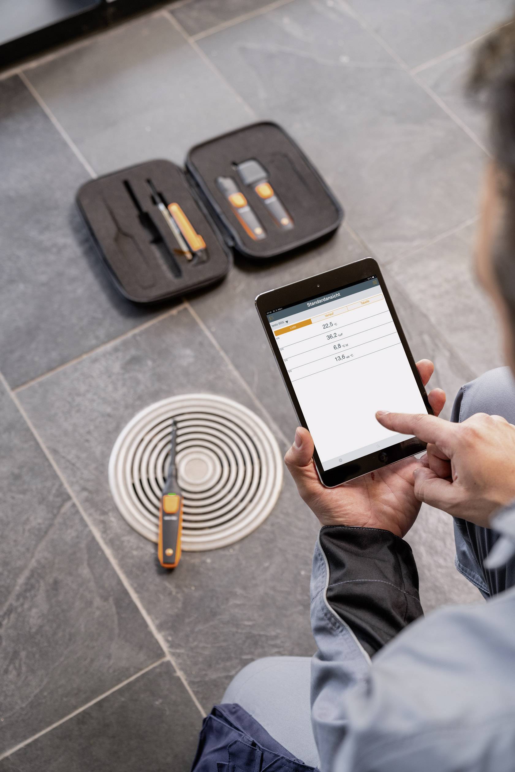 Eine Person bedient ein Tablet zur Messung von Daten, daneben liegt Werkzeug auf einem Bodenablauf.