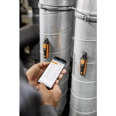 Eine Person verwendet ein Smartphone zur Datenüberwachung an zwei silbernen Rohren mit orangefarbenen Sensoren.