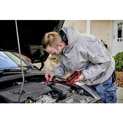'Mann testet Autobatterie mit elektrischem Testgerät bei geöffneter Motorhaube vor einem Haus'