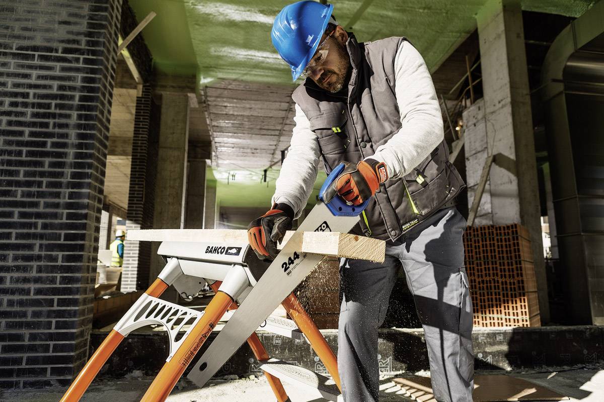 Ein Bauarbeiter mit blauem Helm und Handschuhen sägt ein Holzstück auf einer Baustelle. Im Hintergrund sind Gerüste sichtbar.