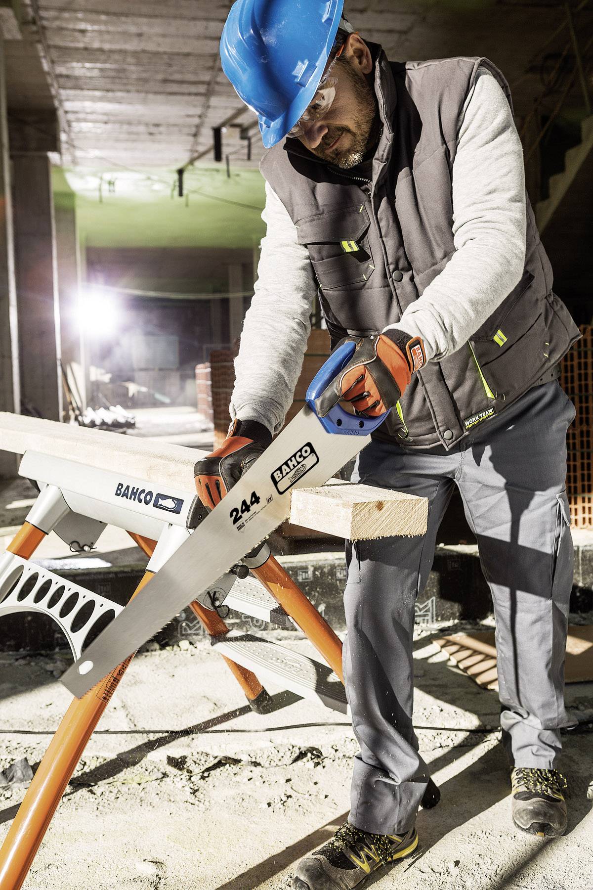 Ein Bauarbeiter mit einem blauen Schutzhelm sägt ein Holzbrett mit einer Handsäge auf einer Baustelle durch.