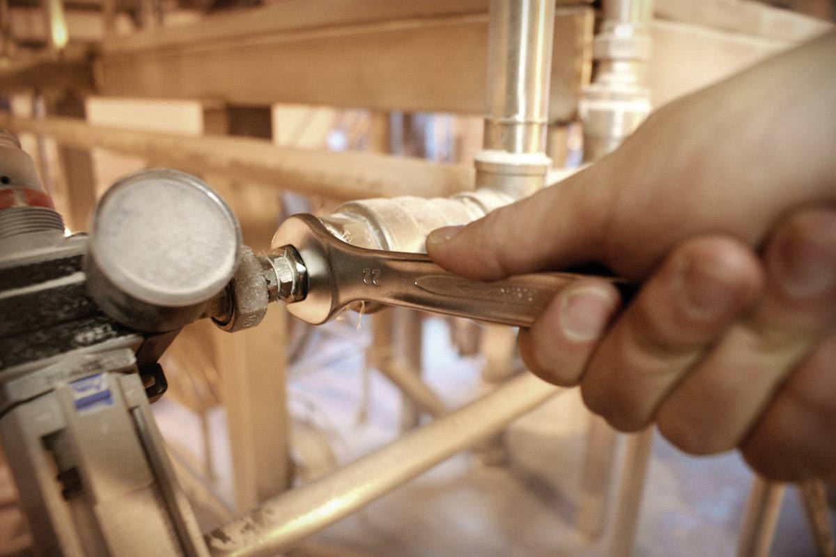 Hand zieht Schraube mit einem Schraubenschlüssel an einer Rohrverbindung fest. Industrielle Umgebung im Hintergrund.
