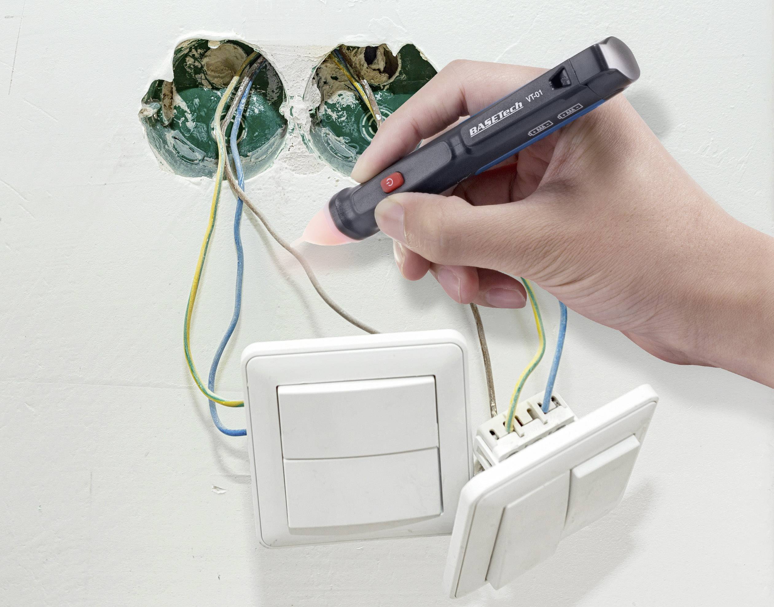 Eine Hand hält einen Spannungsprüfer an freiliegenden Elektrikerkabeln in einer Wand mit zwei Lichtschaltern.