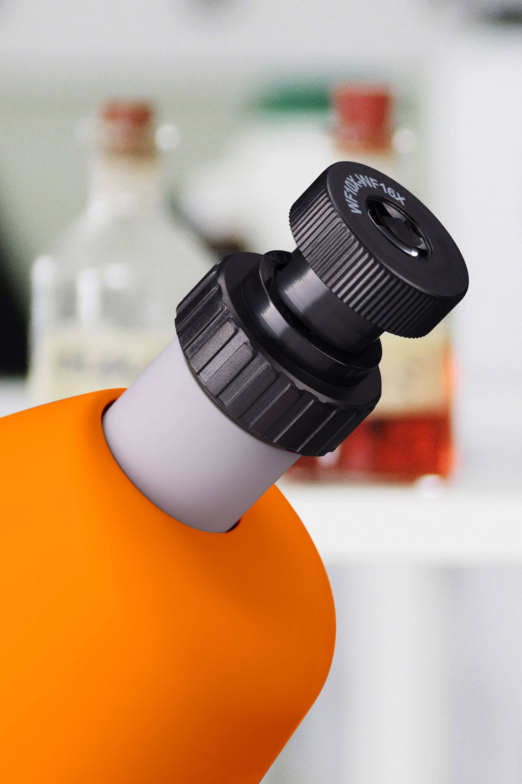 Ein Mikroskopobjektiv in Nahaufnahme fokussiert, mit unscharfen Flaschen im Hintergrund, die eine Laborumgebung andeuten.