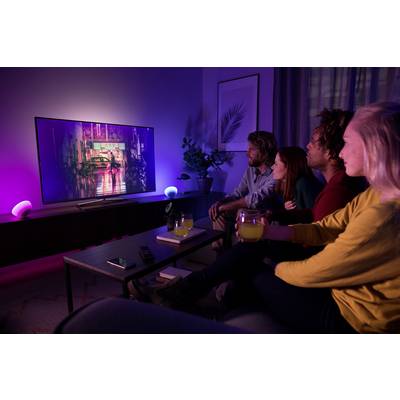 Eine Gruppe von vier Personen sitzt in einem abgedunkelten Wohnzimmer und sieht auf einen großen Fernseher mit bunter Hintergrundbeleuchtung.