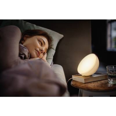Frau liegt im Bett, blickt auf eine leuchtende Lampe auf dem Nachttisch. Atmosphärische Beleuchtung vermittelt Entspannung.