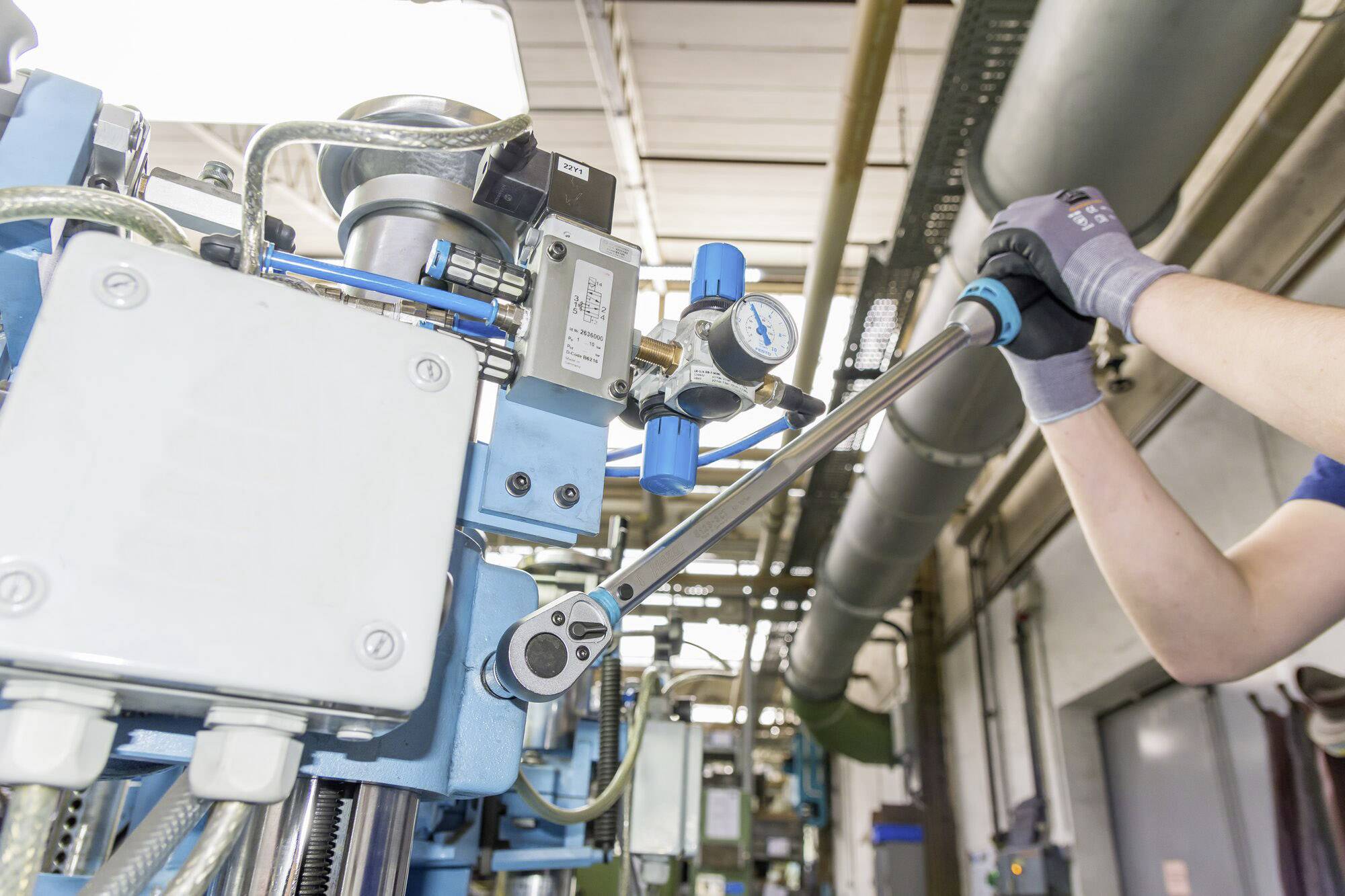 Ein Techniker justiert eine Maschine in einer Fabrikhalle mit einem Drehmomentschlüssel. Rohre und Messgeräte sind im Hintergrund sichtbar.