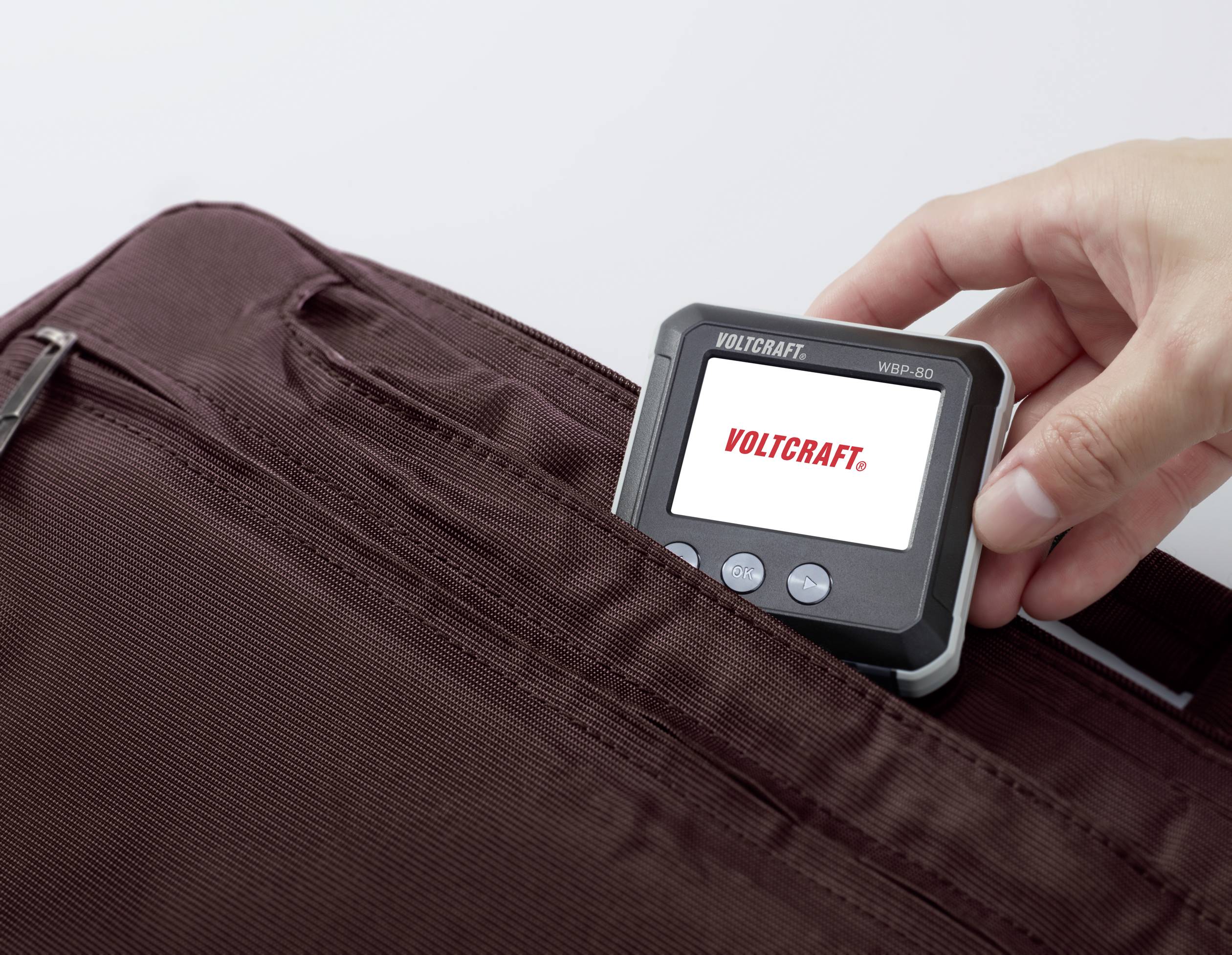 Hand steckt ein digitales Messgerät mit Bildschirm in eine braune Stofftasche. Auf dem Display steht 'VOLTCRAFT'.