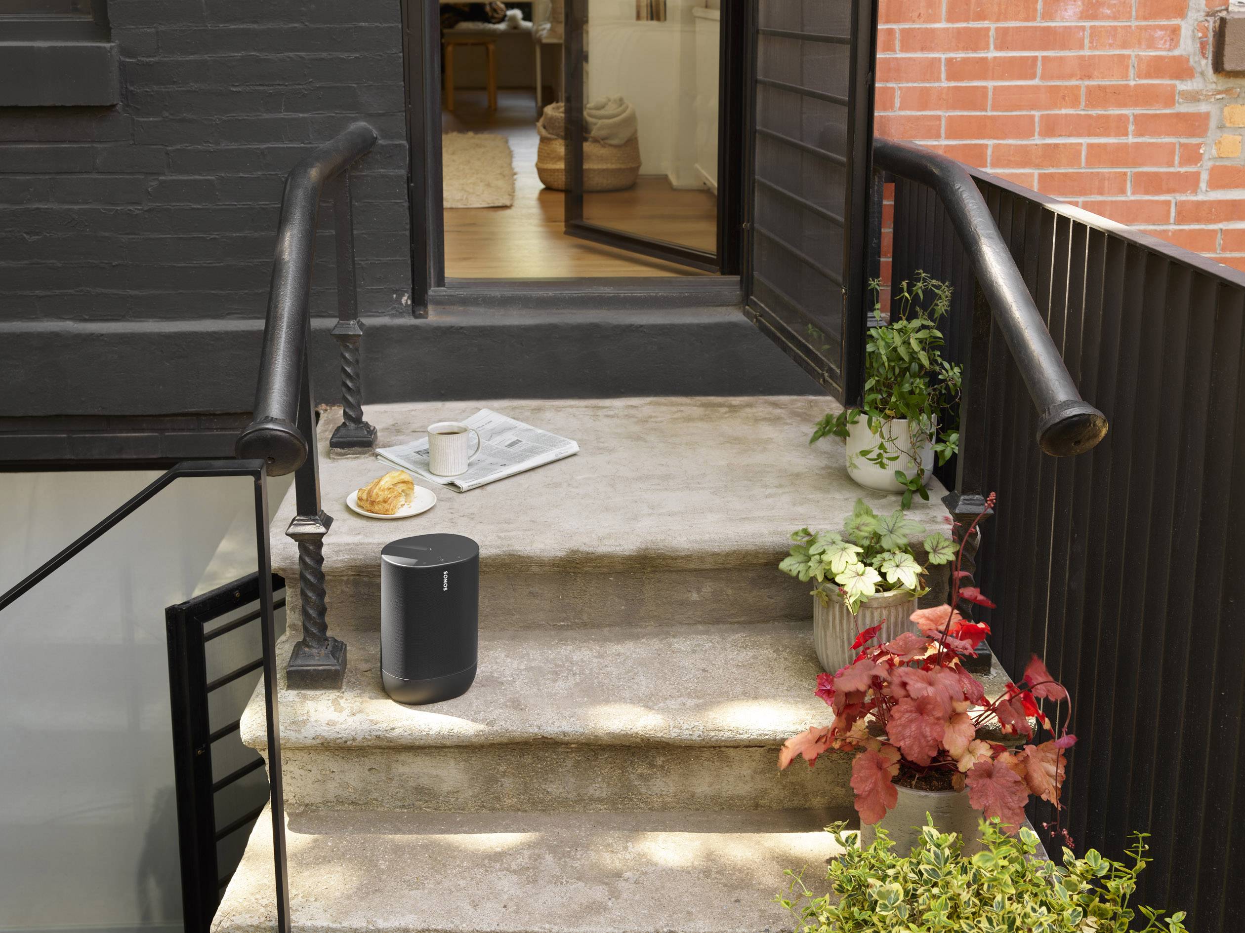 Ein Lautsprecher steht auf einer Treppe neben einer Kaffeetasse und einem Croissant. Pflanzen säumen den Eingang zu einem offenen Raum.