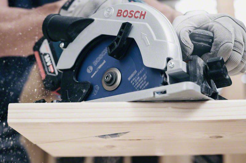 Eine Hand hält eine elektrische Kreissäge mit Bosch-Logo, die ein Holzbrett durchschneidet. Holzspäne fliegen während des Schneidens.