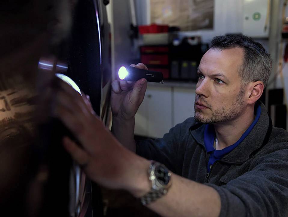 Ein Mann untersucht mit einer Taschenlampe aufmerksam die Oberfläche eines Autos in einer Werkstattumgebung.