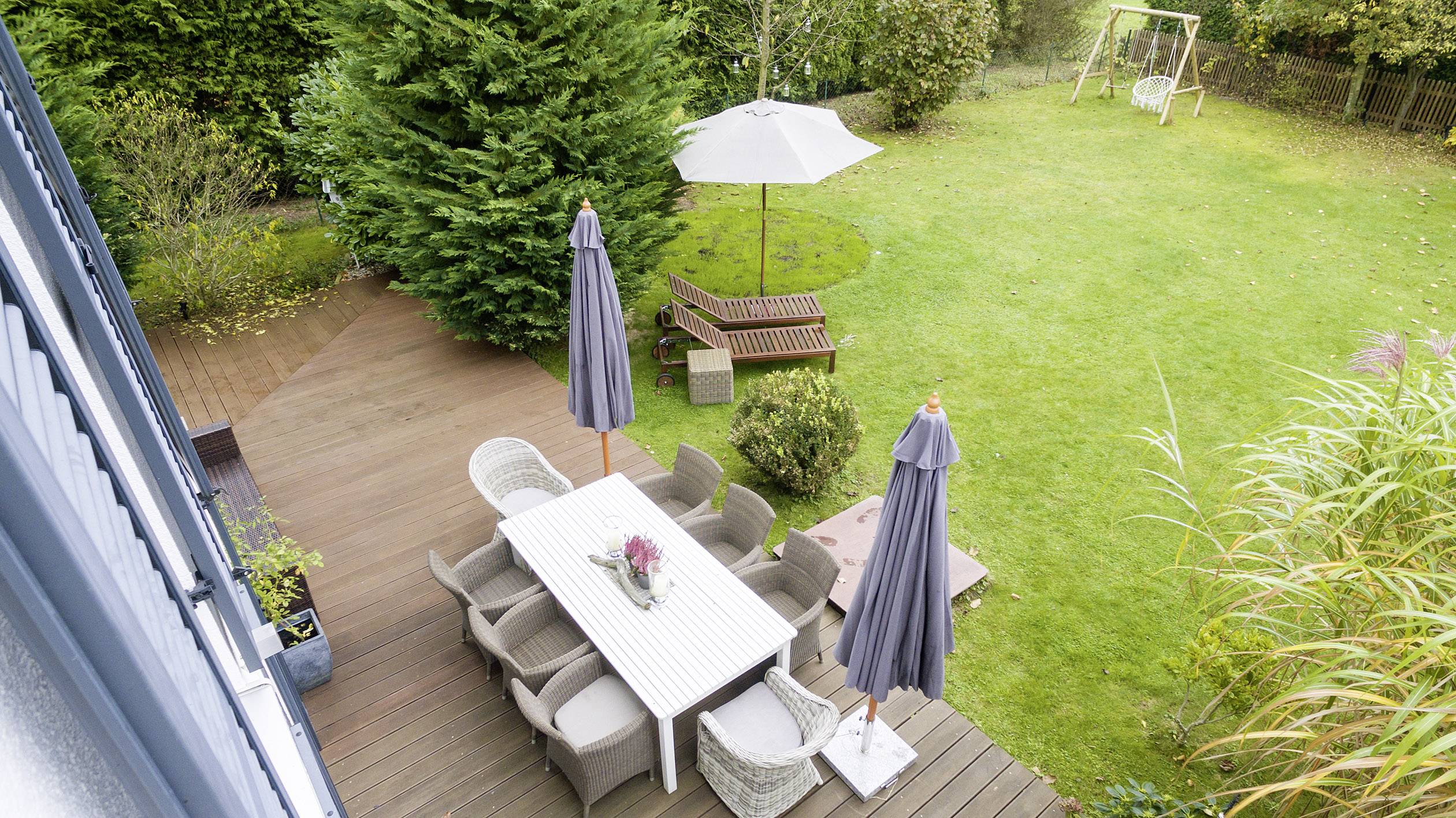 Eine Terrasse mit Gartenmöbeln unter Sonnenschirmen, umgeben von einem grünen Garten mit Rasen und einem Holztisch, im Hintergrund eine Schaukel.