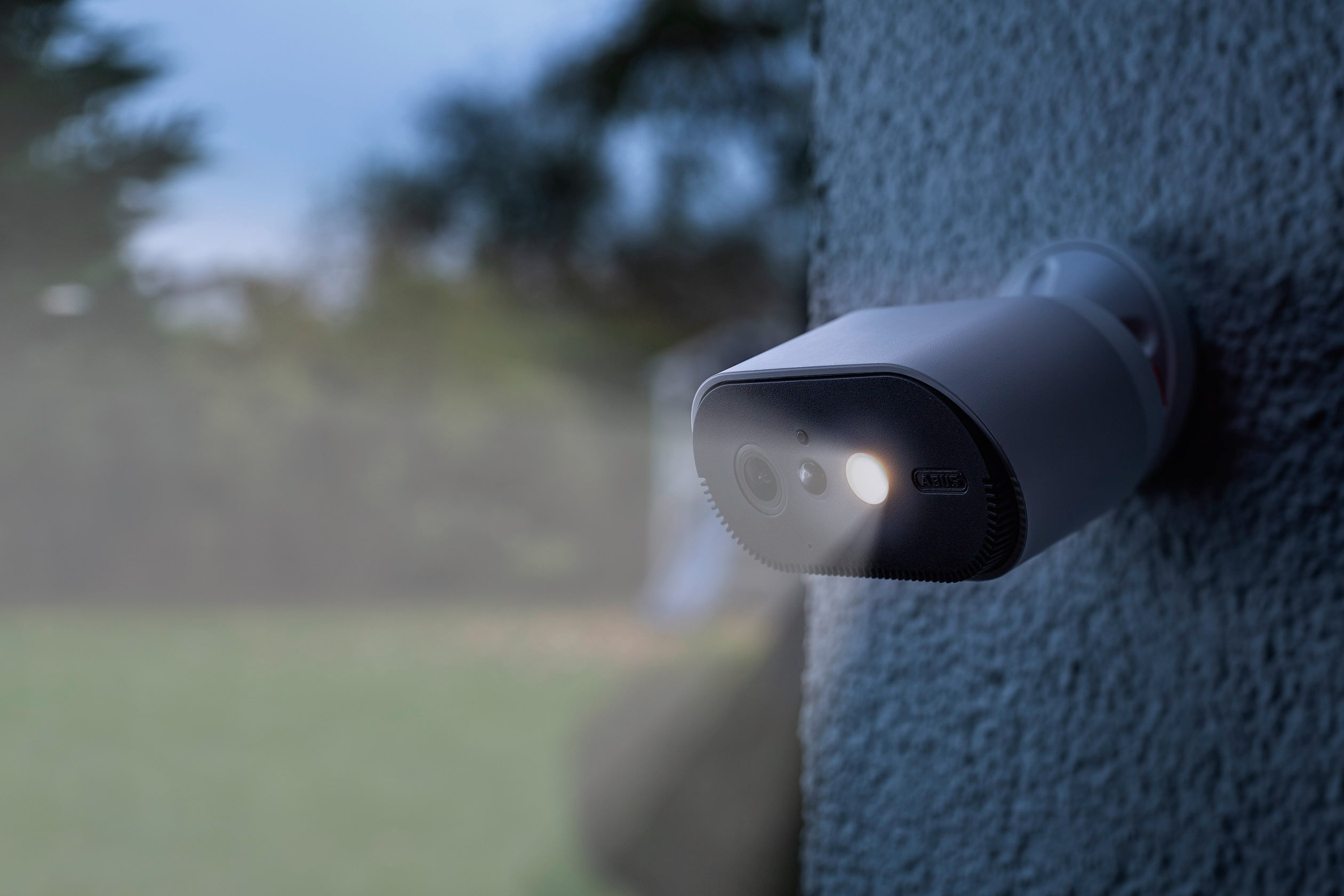 Eine Überwachungskamera mit eingeschaltetem Licht ist außen an einer Gebäudewand montiert und zeigt auf einen Gartenbereich.