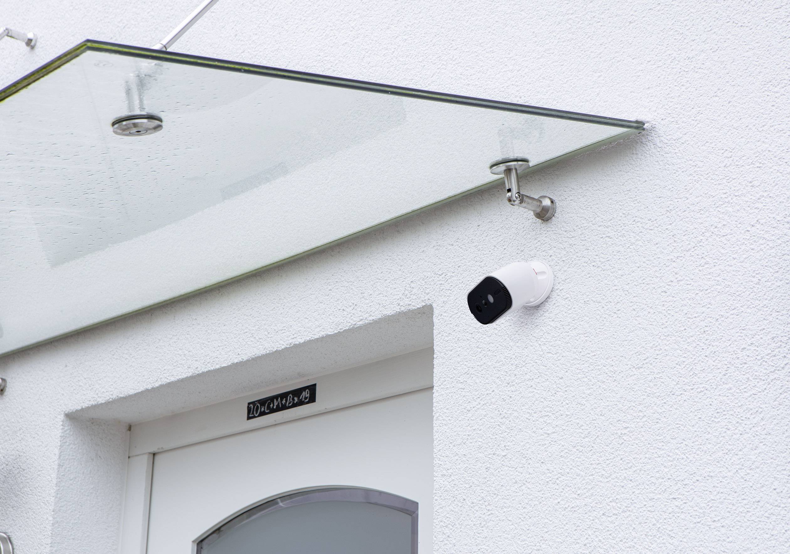 Über einer Haustür mit gläsernem Vordach ist eine Überwachungskamera an der weißen Wand angebracht.