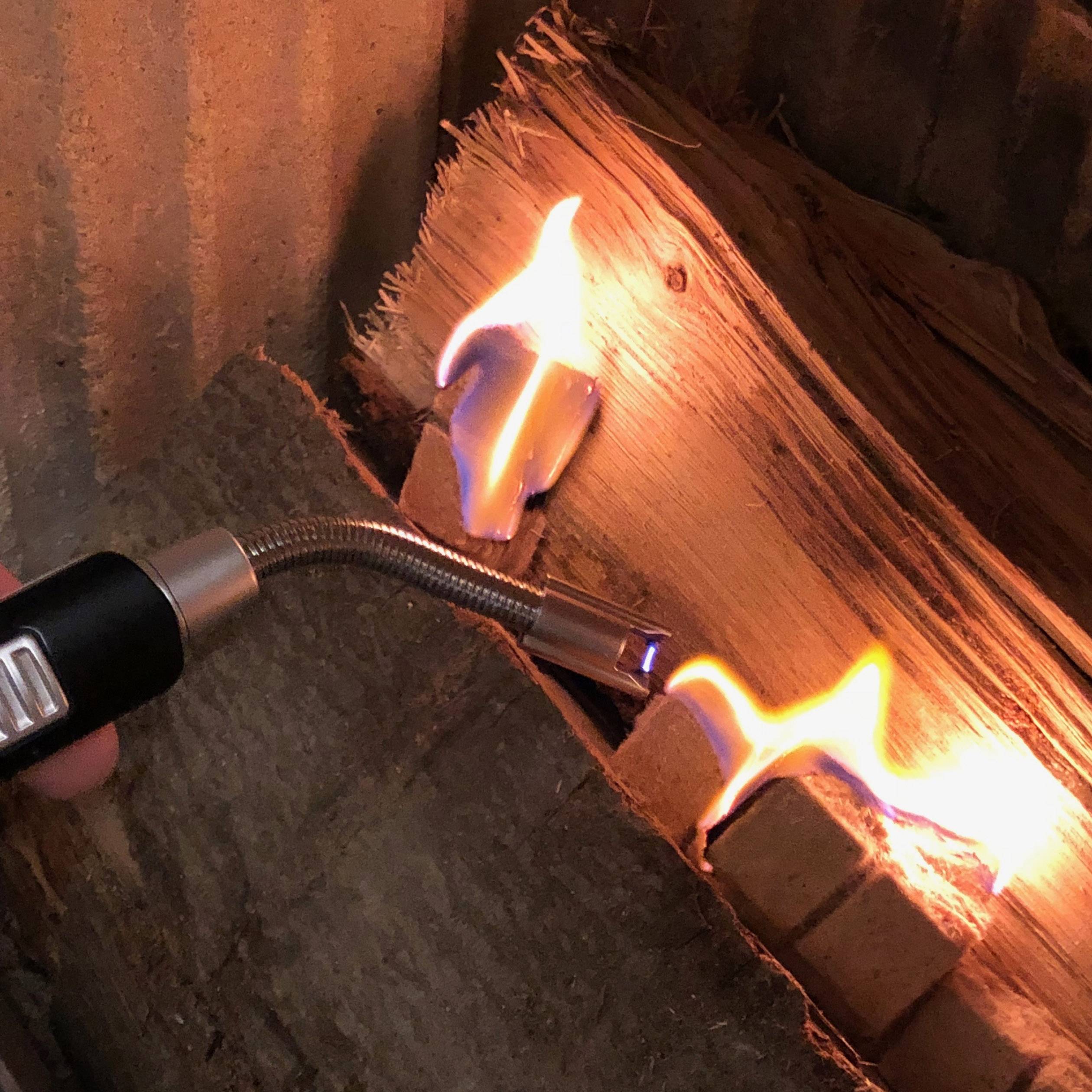 Eine Hand hält ein Gasbrenner und entzündet Holzscheite in einem Kamin. Flammen leuchten hell.