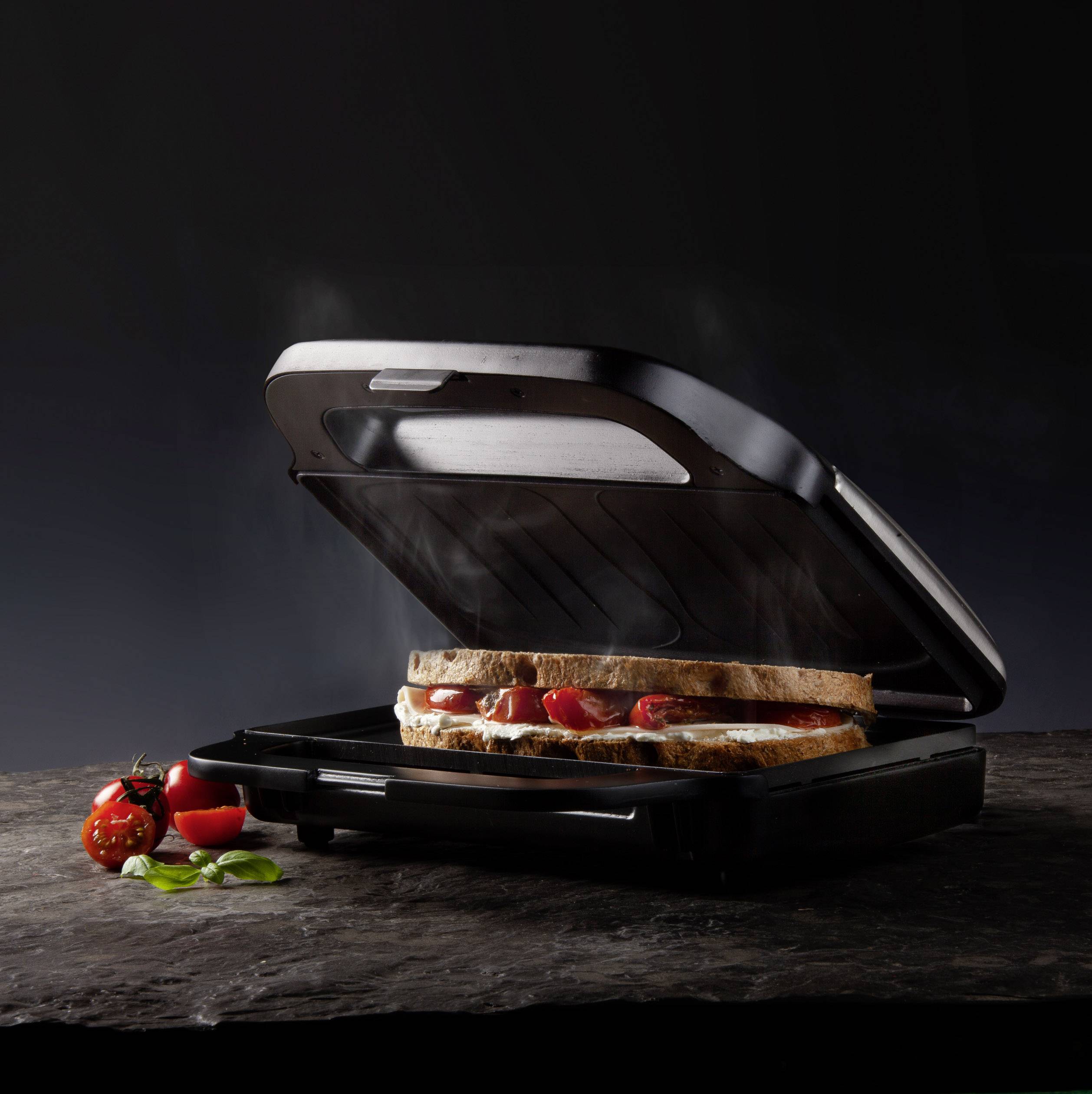 Ein Sandwichmaker auf dunklem Hintergrund, grillt ein belegtes Sandwich. Daneben liegen frische Tomaten und Basilikumblätter.