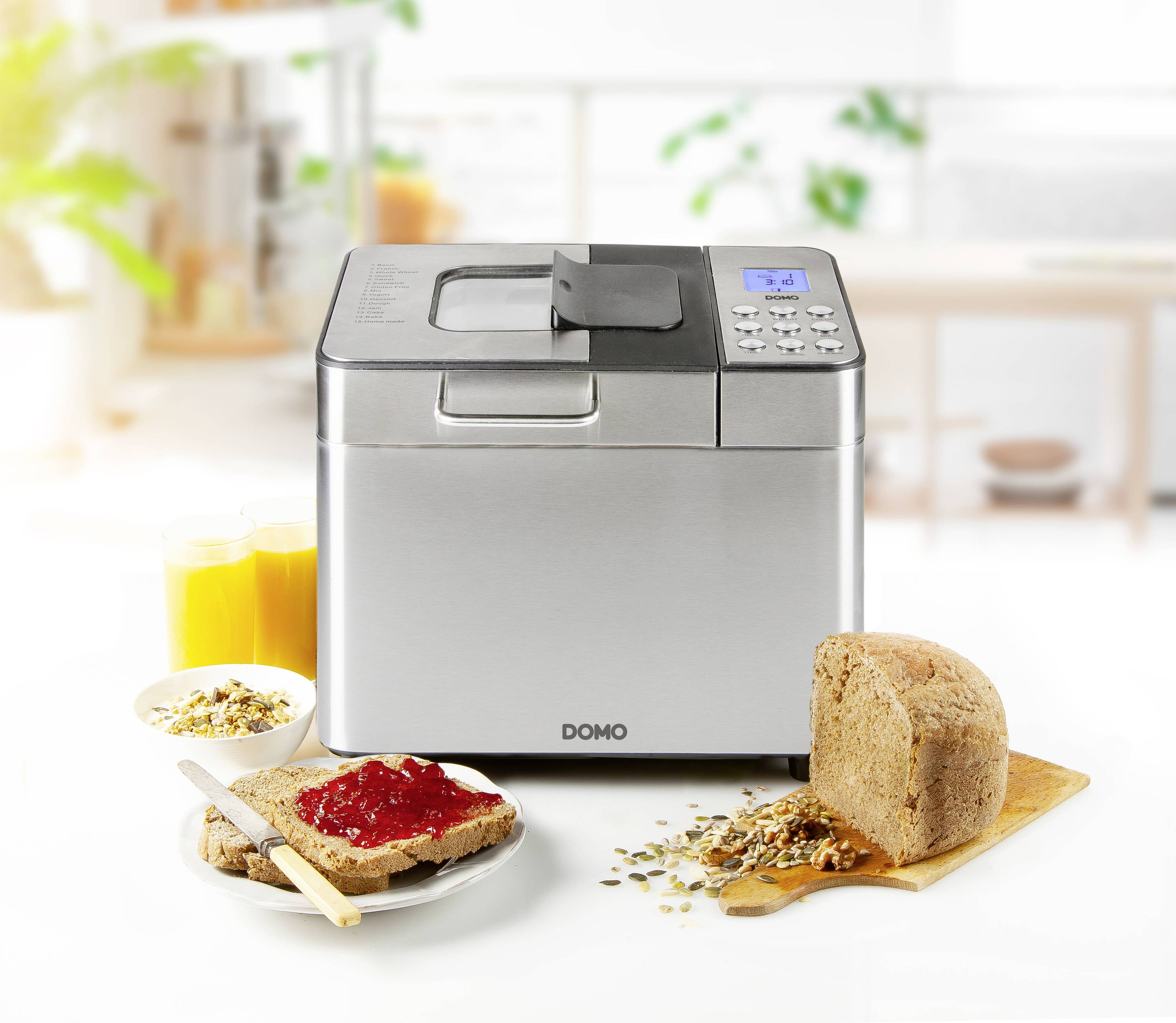 Brotbackautomat auf einer Küchenzeile mit gebackenem Brot auf einem Holzbrett, Müsli, Toast mit Marmelade und zwei Gläsern Orangensaft.