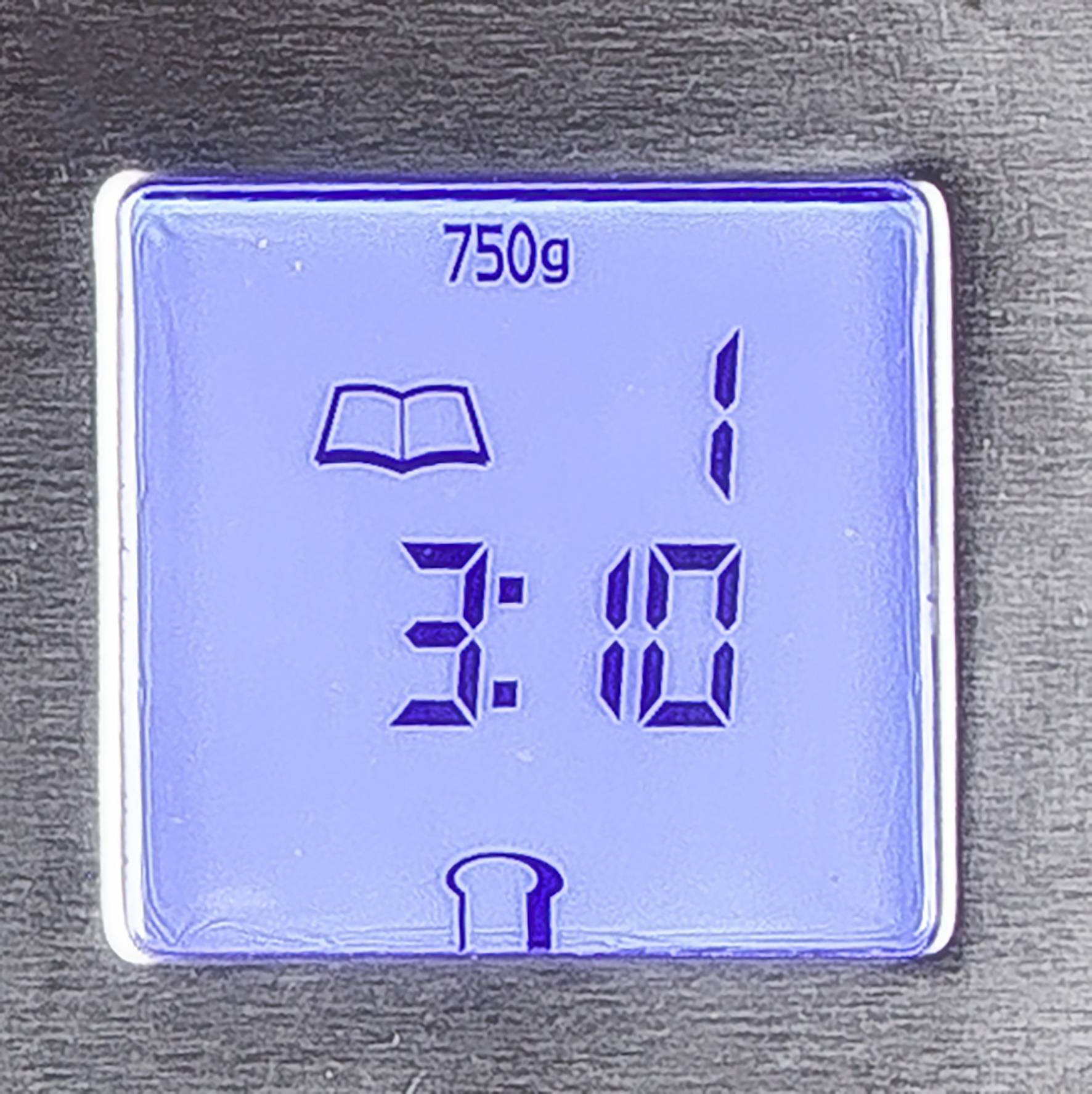 Digitalanzeige eines kleinen Bildschirmes auf einem Gerät. Zeigt oben '750g', darunter ein Buchsymbol und daneben '1'. Unten Uhrzeit '3:10' und ein Toastsymbol.