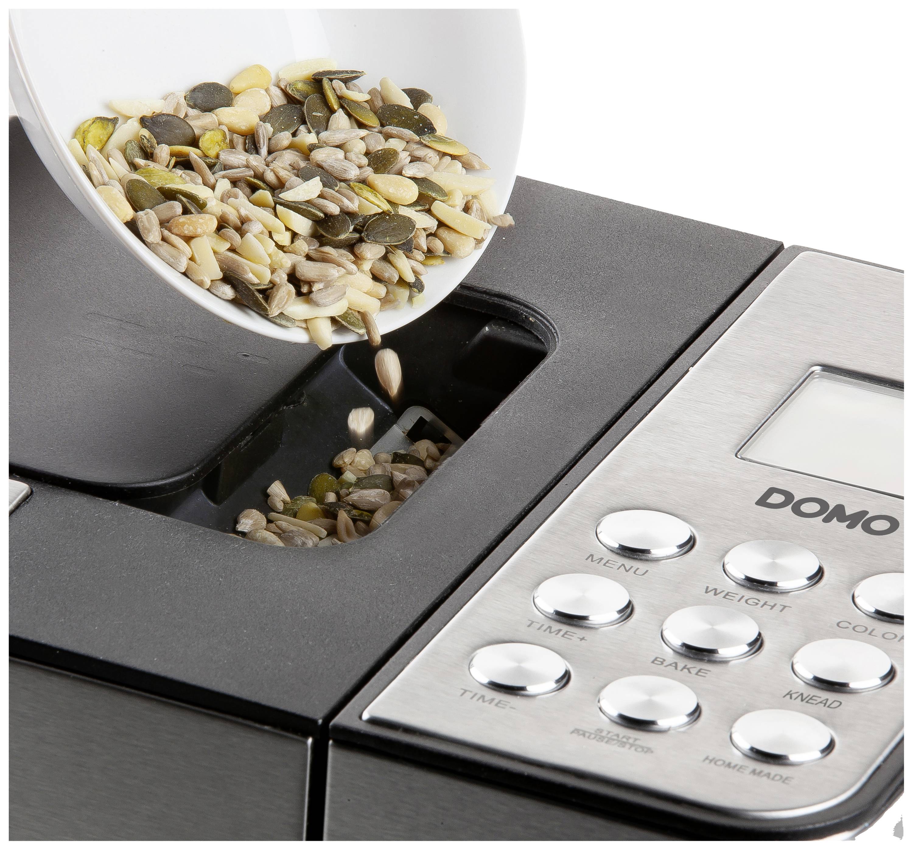 Eine Brotbackmaschine, in die Körner aus einer Schüssel in den Zutatenbehälter gefüllt werden. Die Maschine hat ein Bedienfeld.