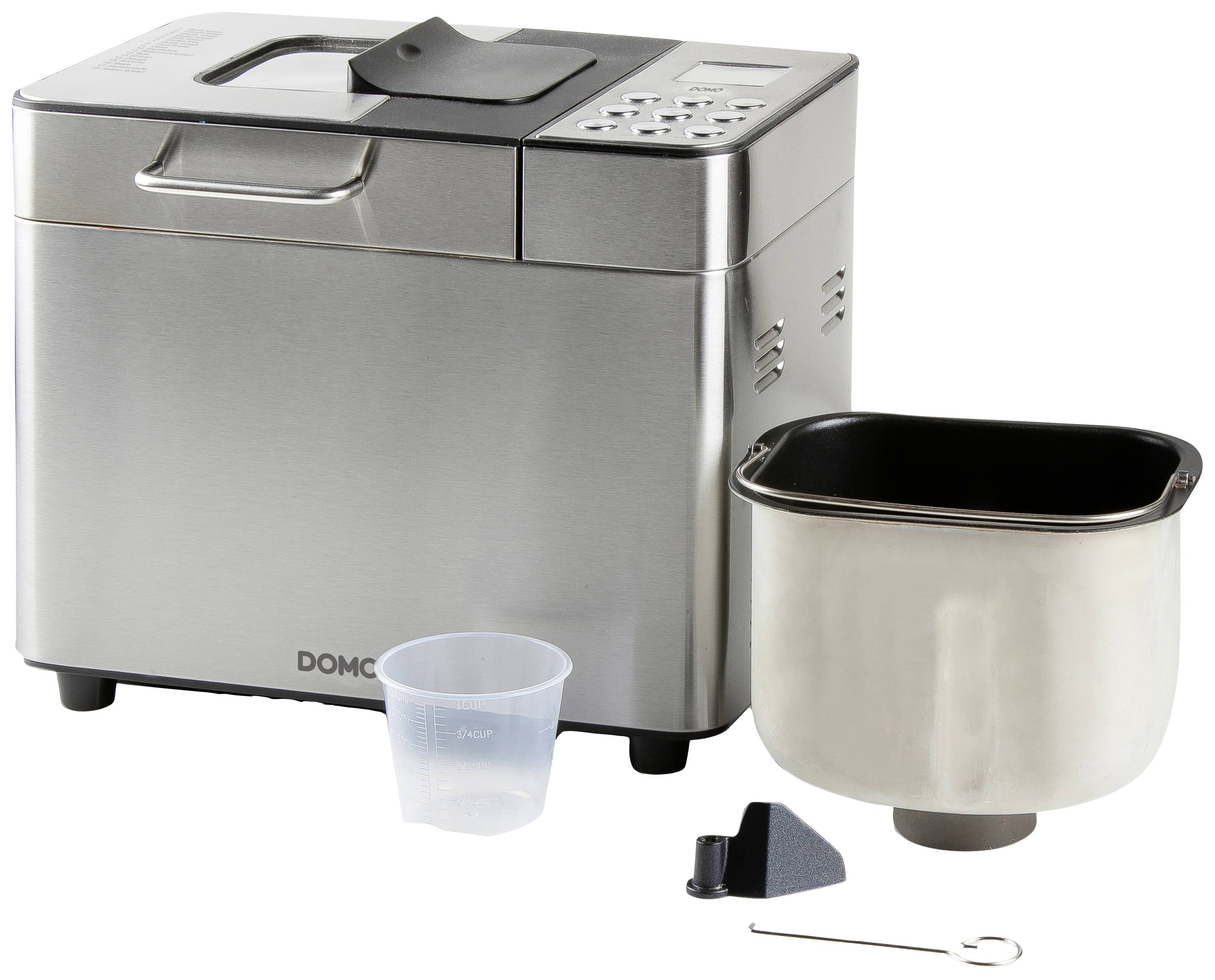 Brotbackautomat mit silbernem Gehäuse, herausnehmbarer Backform, Messbecher, Knethaken und einem Zubehörteil auf einer weißen Fläche.