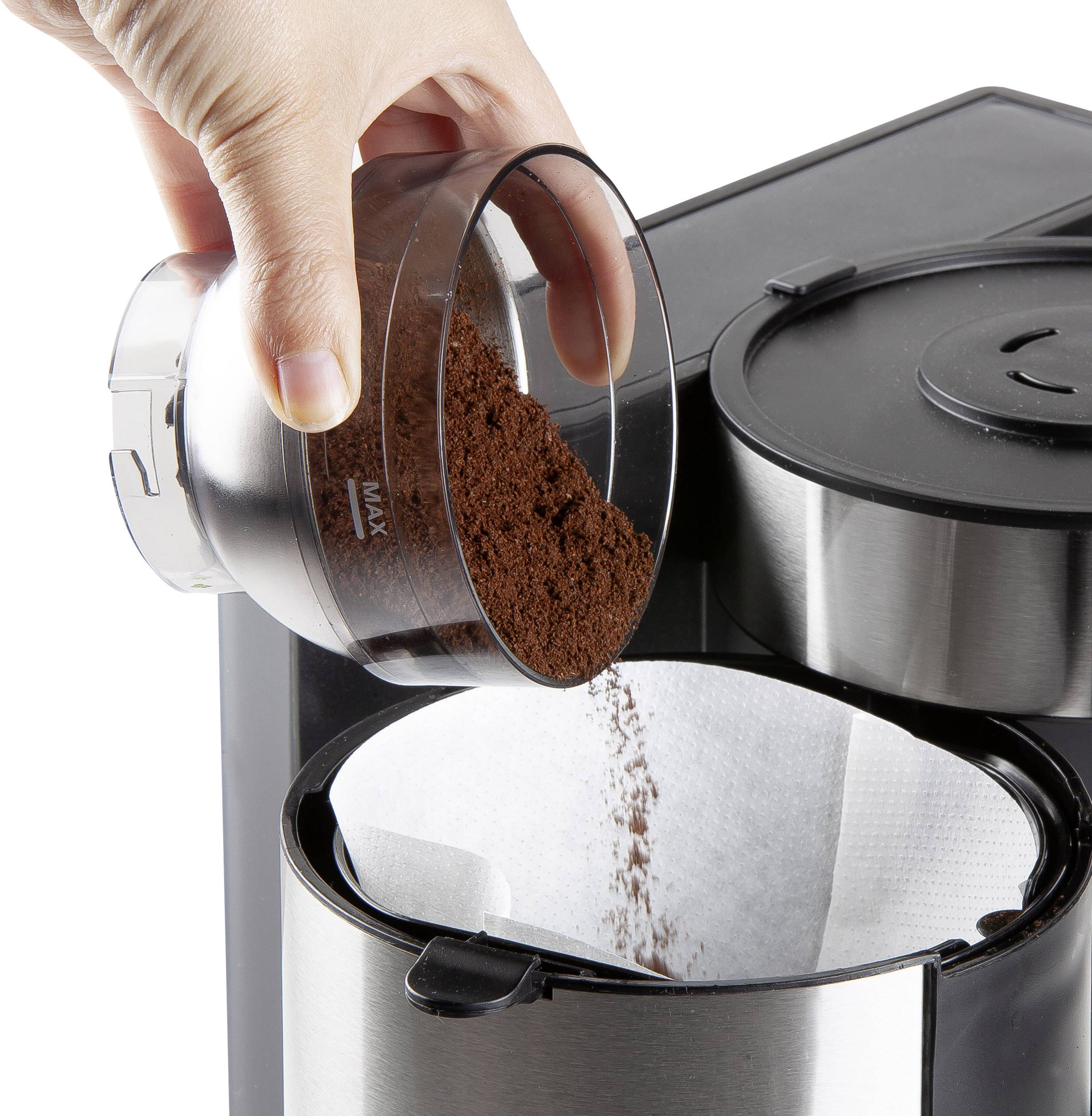 Hand füllt Kaffeepulver in eine Filtermaschine. Der Filter ist oben geöffnet, und die Maschine ist aus Edelstahl und schwarz.