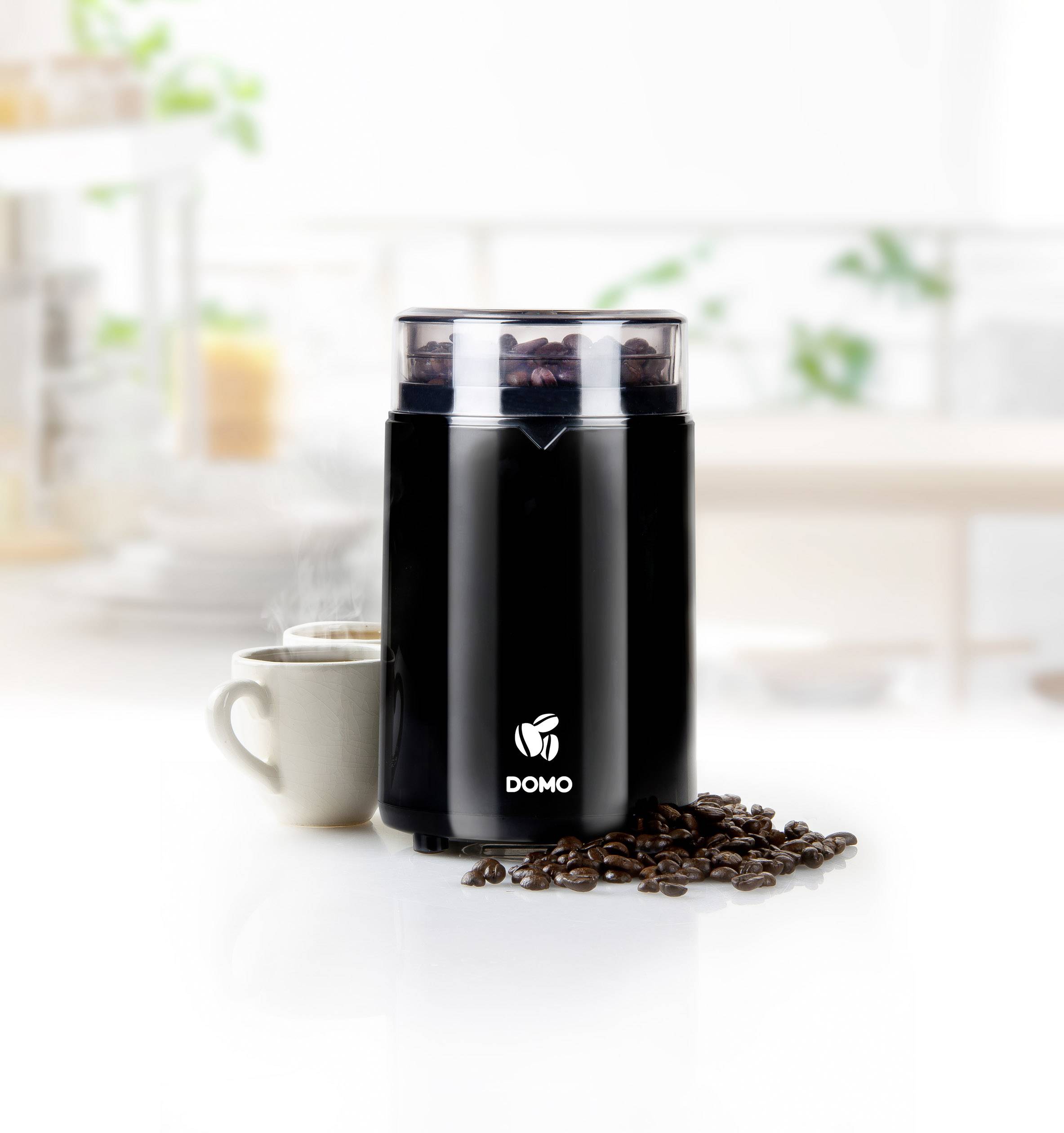 Eine schwarze Kaffeemühle mit dem Logo 'DOMO', umgeben von Kaffeebohnen, neben einer Tasse Kaffee auf einer Küchentheke.
