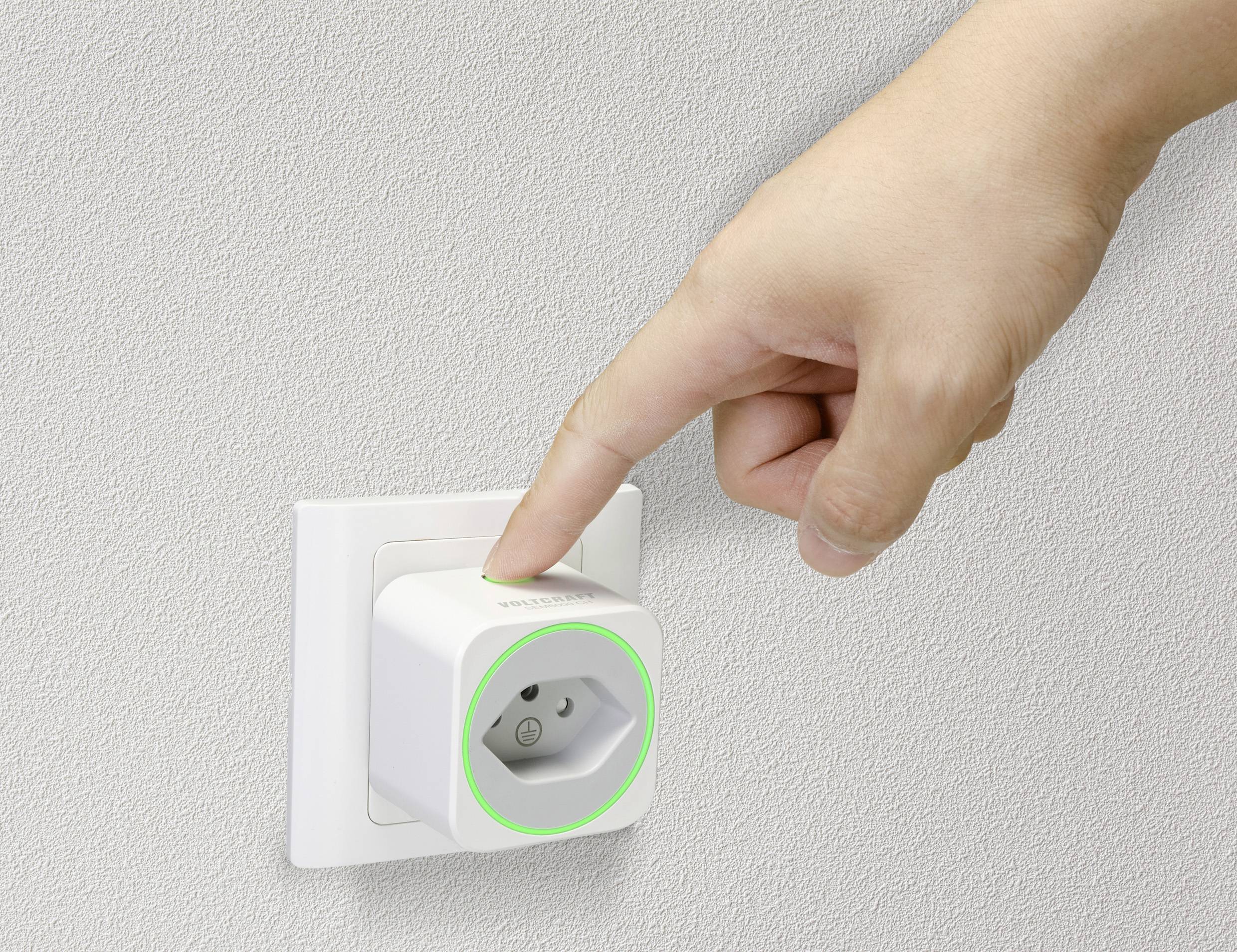 'Eine Hand steckt einen smarten Stecker in eine weiße Wandsteckdose, der Stecker leuchtet grün. Der Hintergrund ist beige.'