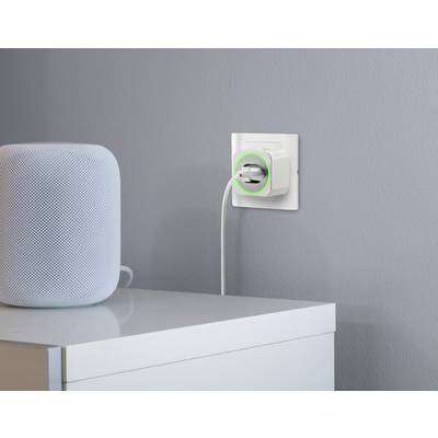 Weiße Steckdose an grauer Wand, ein Smart-Home-Gerät mit grünem Leuchtring. Daneben auf einem weißen Tisch steht ein Lautsprecher.
