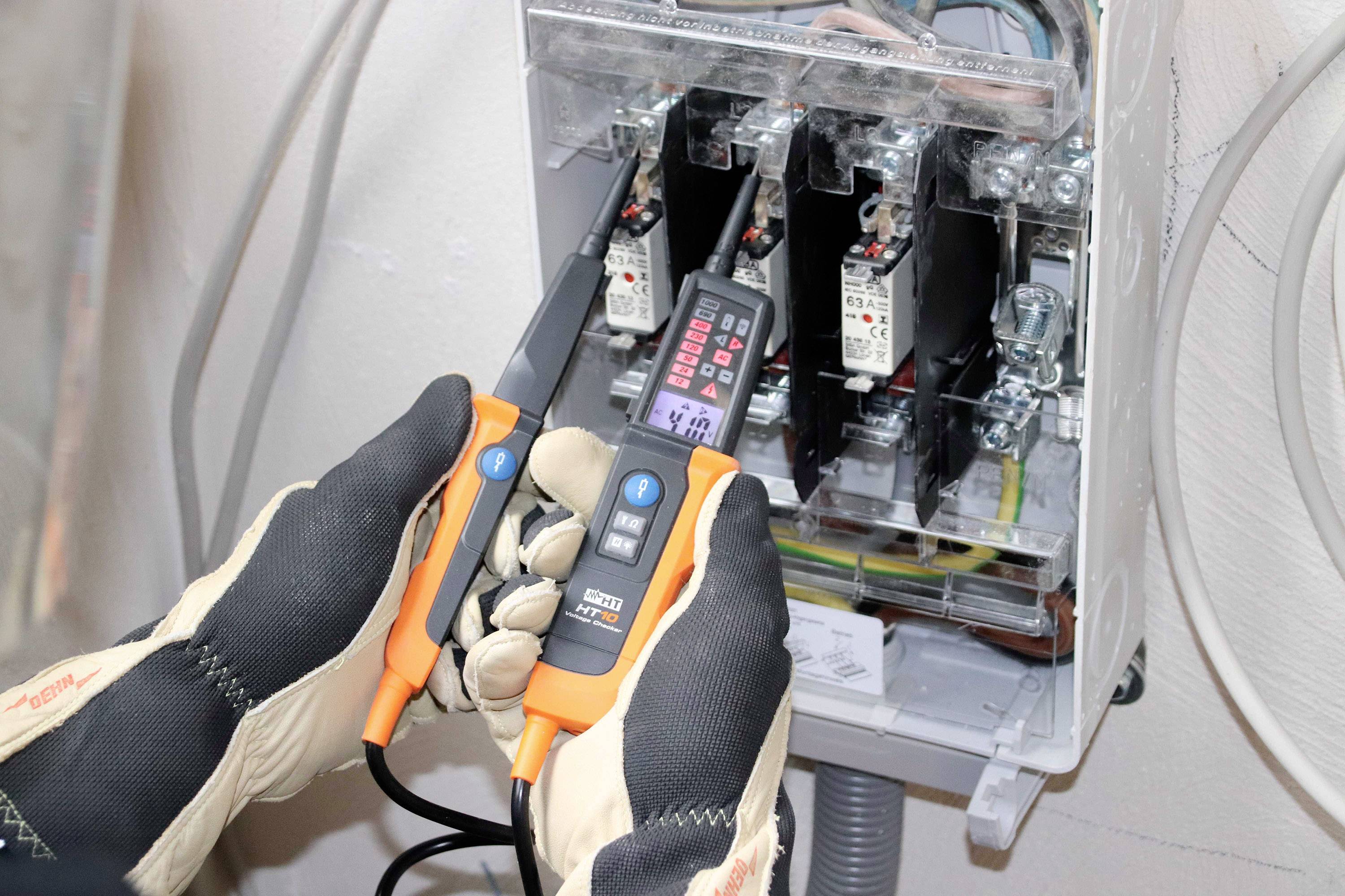 'Elektriker trägt Handschuhe und verwendet ein Spannungsprüfgerät an einem offenen Sicherungskasten.'