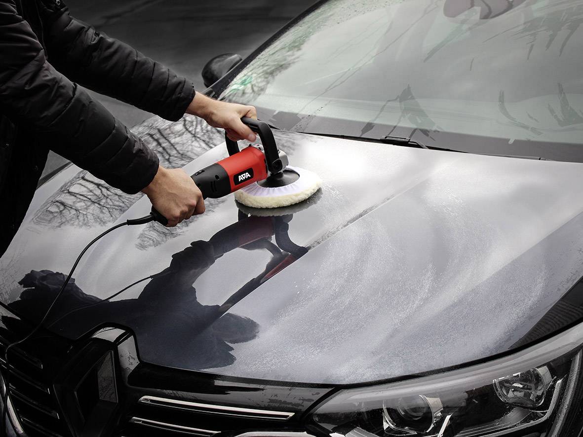 Person poliert mit einer rotierenden Poliermaschine die Motorhaube eines Autos. Reflexionen und benetzte Oberfläche sind sichtbar.