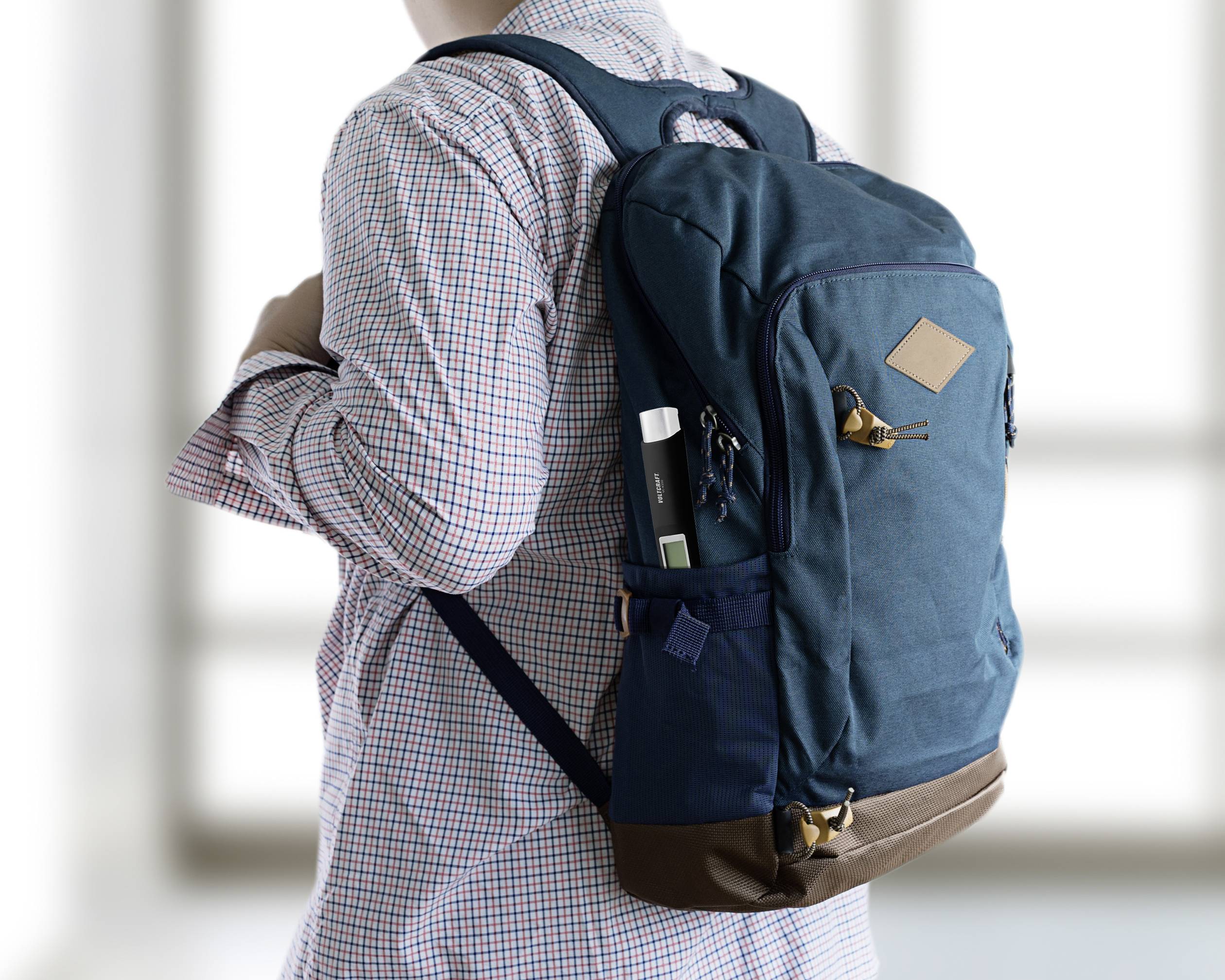 Eine Person trägt einen blauen Rucksack. Der Rucksack hat mehrere Taschen und braune Akzente. Fensterscheiben im Hintergrund.