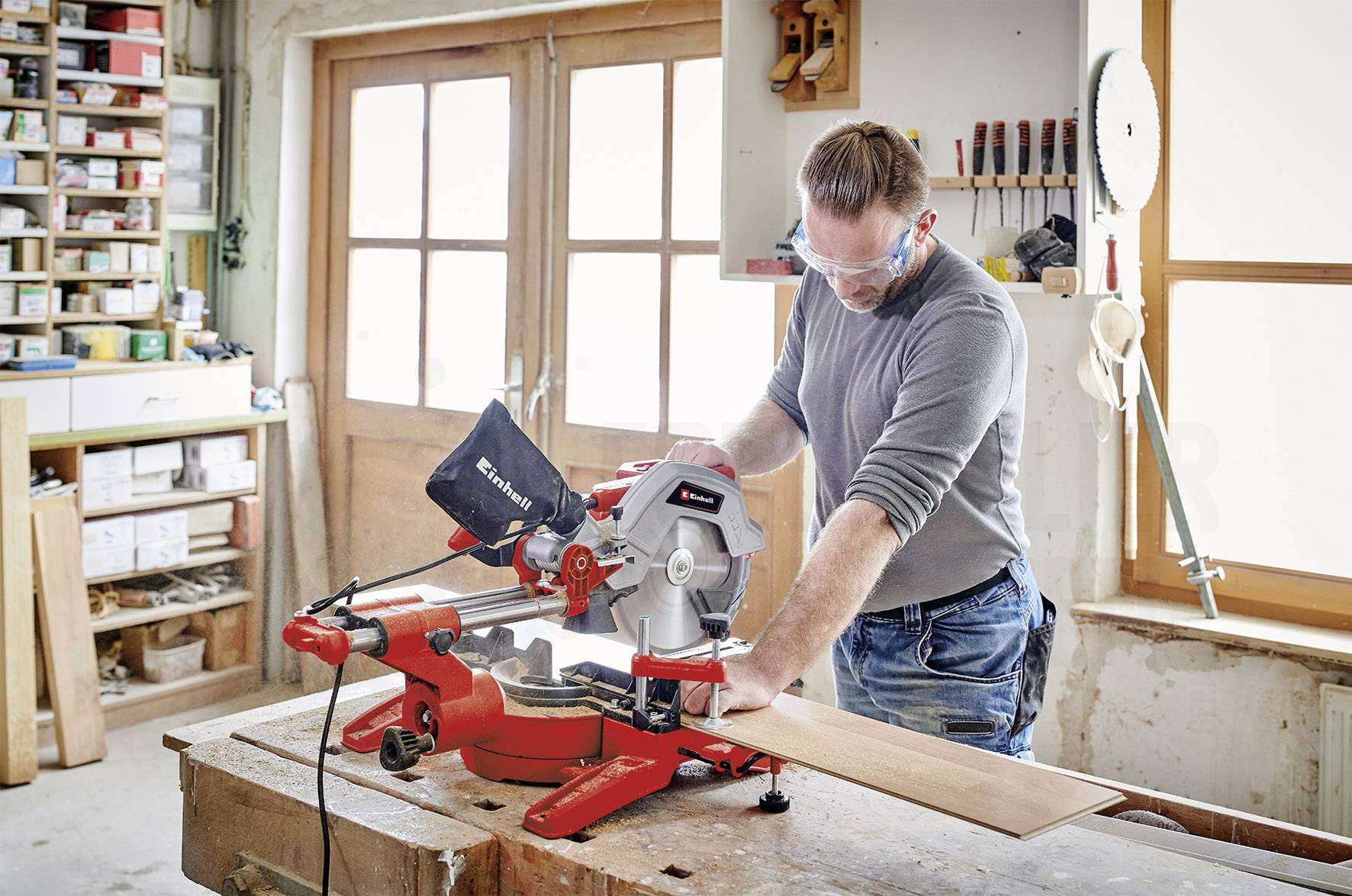 Ein Mann arbeitet in einer Werkstatt mit einer Tischkreissäge, trägt Sicherheitsbrille, Holzstück wird geschnitten.
