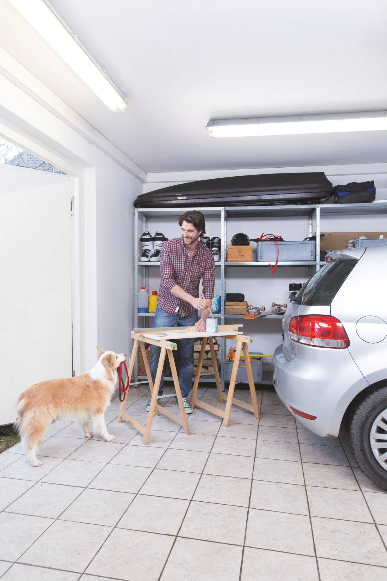 Ein Mann arbeitet an einem Tisch in einer Garage neben einem Auto. Ein Hund steht auf dem Boden und blickt zu ihm auf. Im Hintergrund sind Regale mit Werkzeugen.