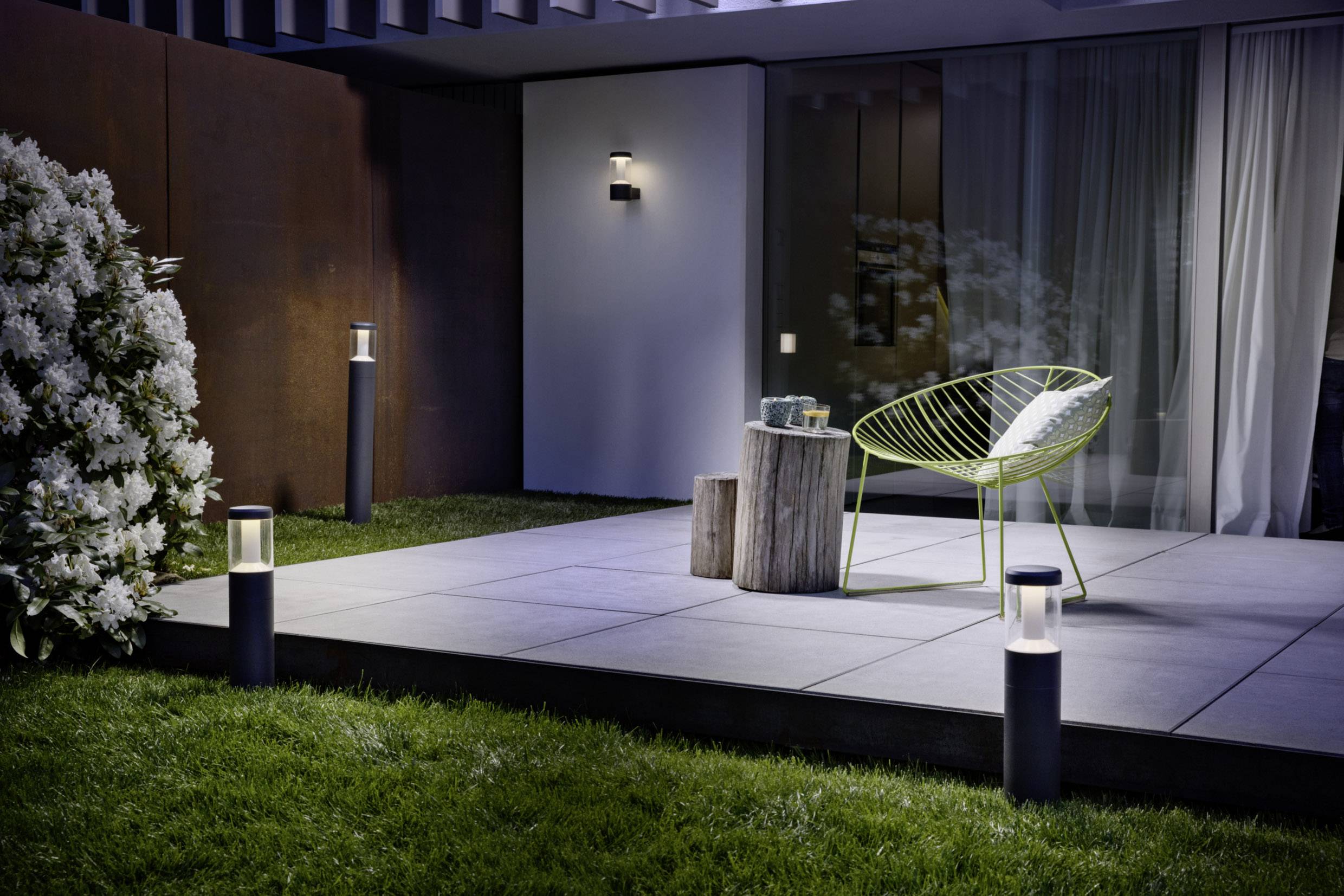 Eine Terrasse bei Nacht, beleuchtet von modernen Außenlampen. Ein grüner Stuhl und Beistelltisch aus Holz stehen auf den Fliesen.