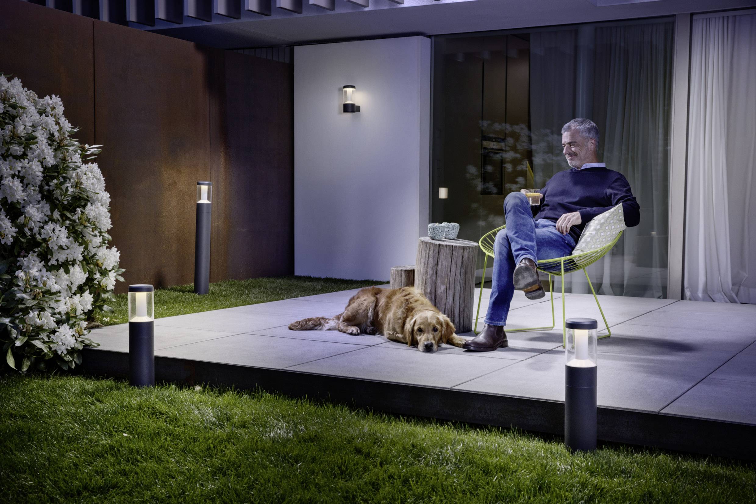 Ein Mann sitzt nachts auf einer Terrasse und hält eine Tasse. Neben ihm liegt ein Hund. Die Beleuchtung sorgt für eine entspannte Atmosphäre.