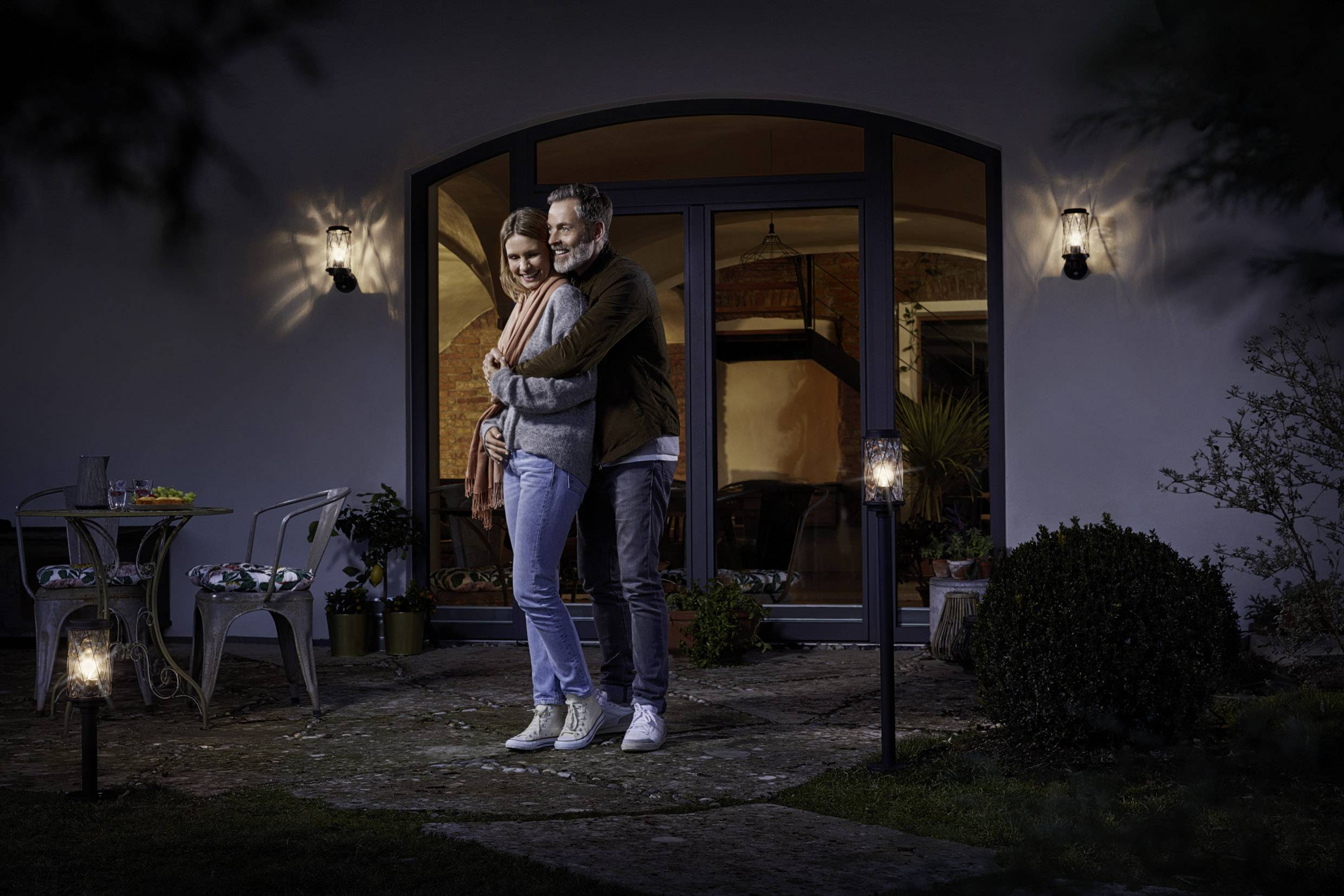 Ein Paar umarmt sich liebevoll im Garten eines Hauses bei Nacht, umgeben von dezenten Außenlaternen und Pflanzen, ein Gefühl von Geborgenheit vermittelnd.