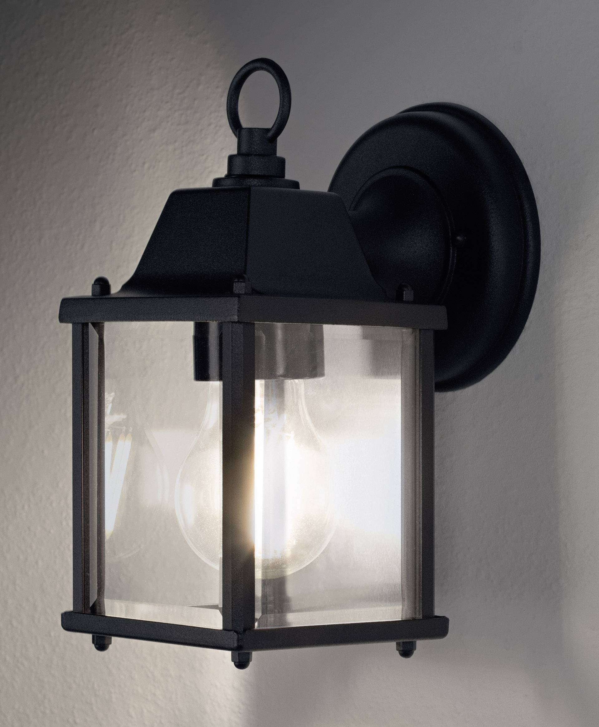 Schwarze Wandleuchte mit transparentem Glas und sichtbarer Glühbirne, montiert an einer hellen Wand, leuchtet sanft.