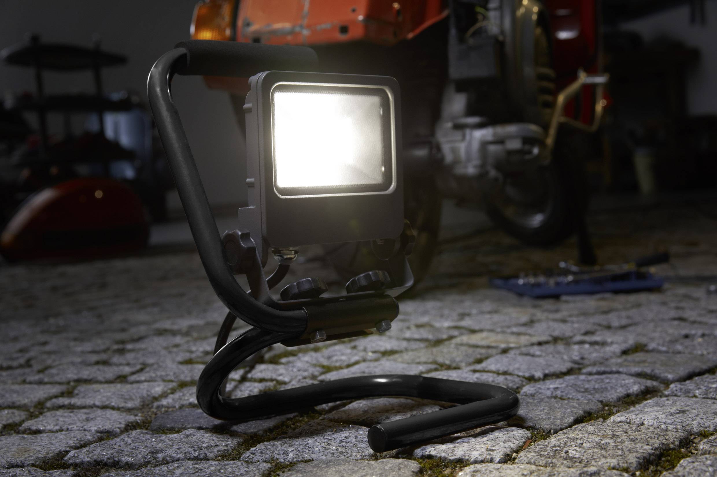 Ein Baustrahler beleuchtet eine gepflasterte Fläche in einer Werkstatt. Im Hintergrund ist unscharf eine Maschine zu sehen.