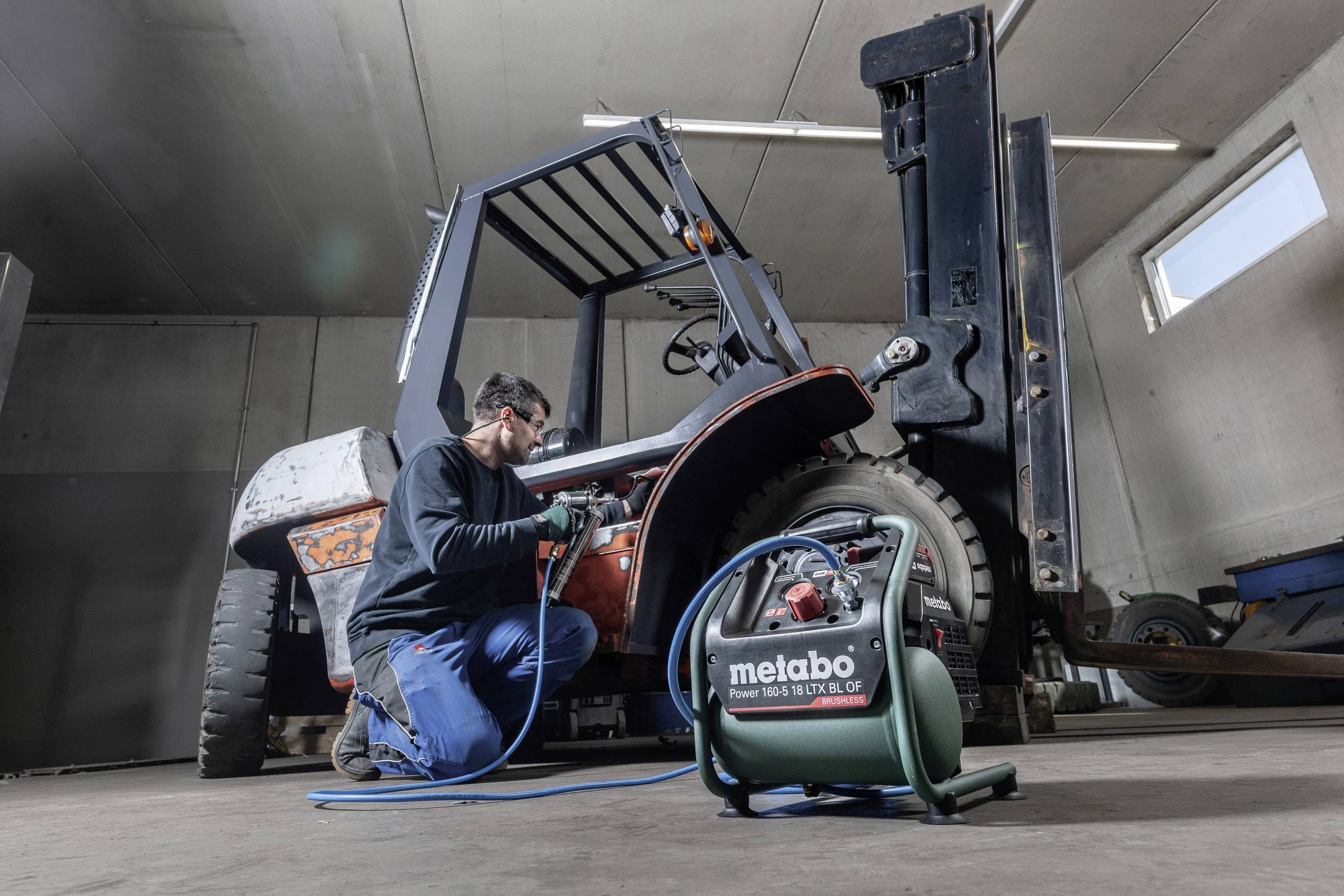 Mechaniker repariert Gabelstapler mit Druckluftwerkzeug; Metabo-Kompressor sichtbar. Werkstattumgebung mit Betonwänden und Schläuchen.