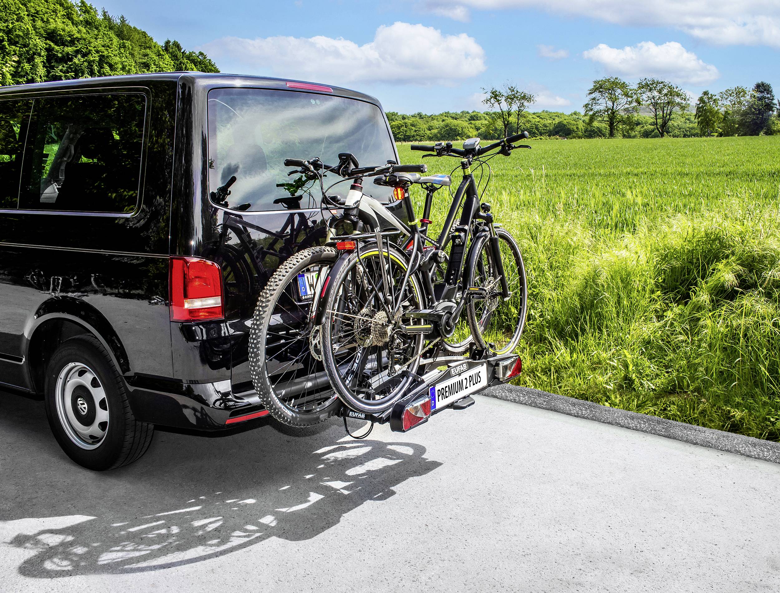 Ein schwarzer Van parkt auf einer Straße, mit zwei Fahrrädern auf einem Träger am Heck, im Hintergrund grüne Felder und Bäume.