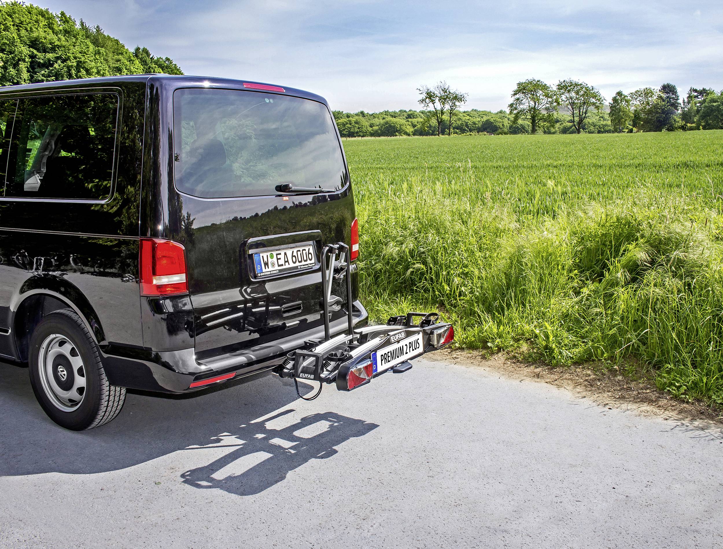 Schwarzer Lieferwagen mit einem Fahrradträger auf einer ländlichen Straße. Im Hintergrund grüne Felder und Bäume unter blauem Himmel.