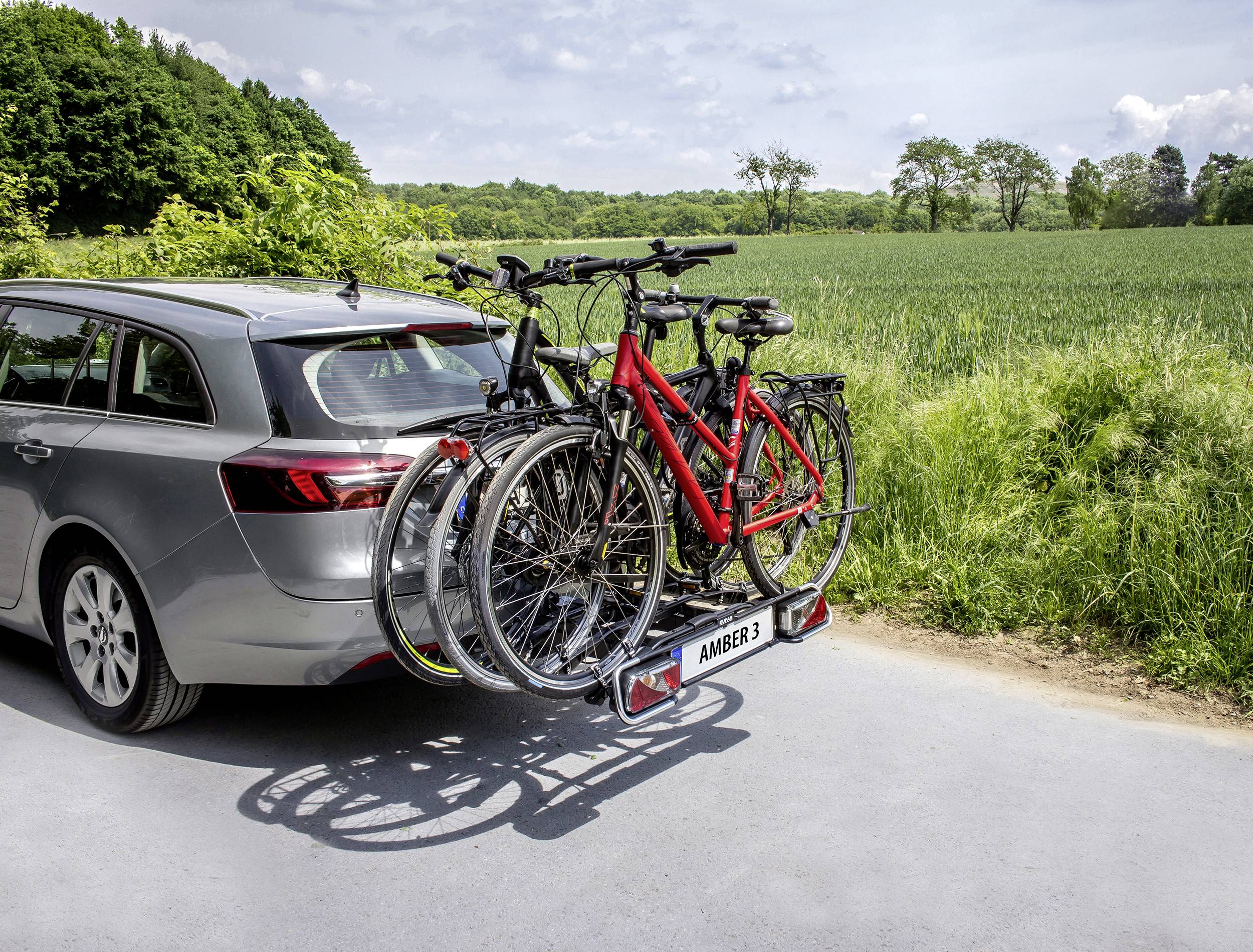 Silbernes Auto mit Fahrradträger am Heck, der zwei Fahrräder transportiert, auf einer Landstraße neben einem grünen Feld.