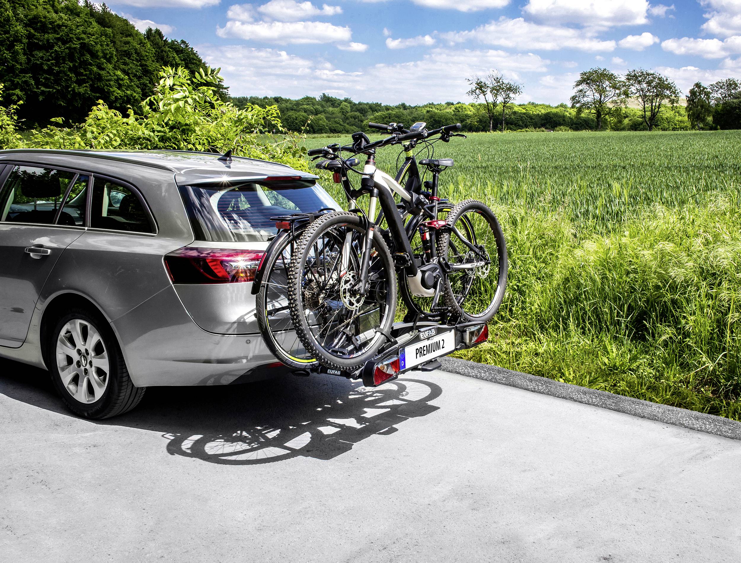 Ein silbernes Auto mit einem Fahrradträger, der zwei Fahrräder trägt, steht auf einem ländlichen Weg neben grünen Feldern und Bäumen.