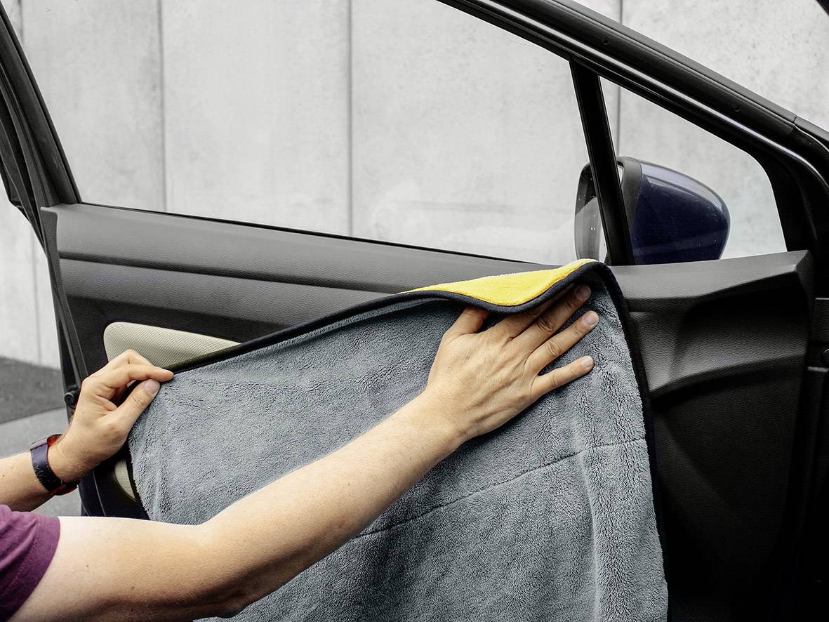 Eine Person reinigt die Innenseite einer Autotür mit einem grauen Handtuch. Im Hintergrund ist eine Betonwand zu sehen.