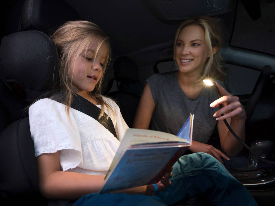 Ein Kind liest ein Buch im Auto, während eine erwachsene Person mit einer kleinen Lampe lächelt und das Buch beleuchtet.