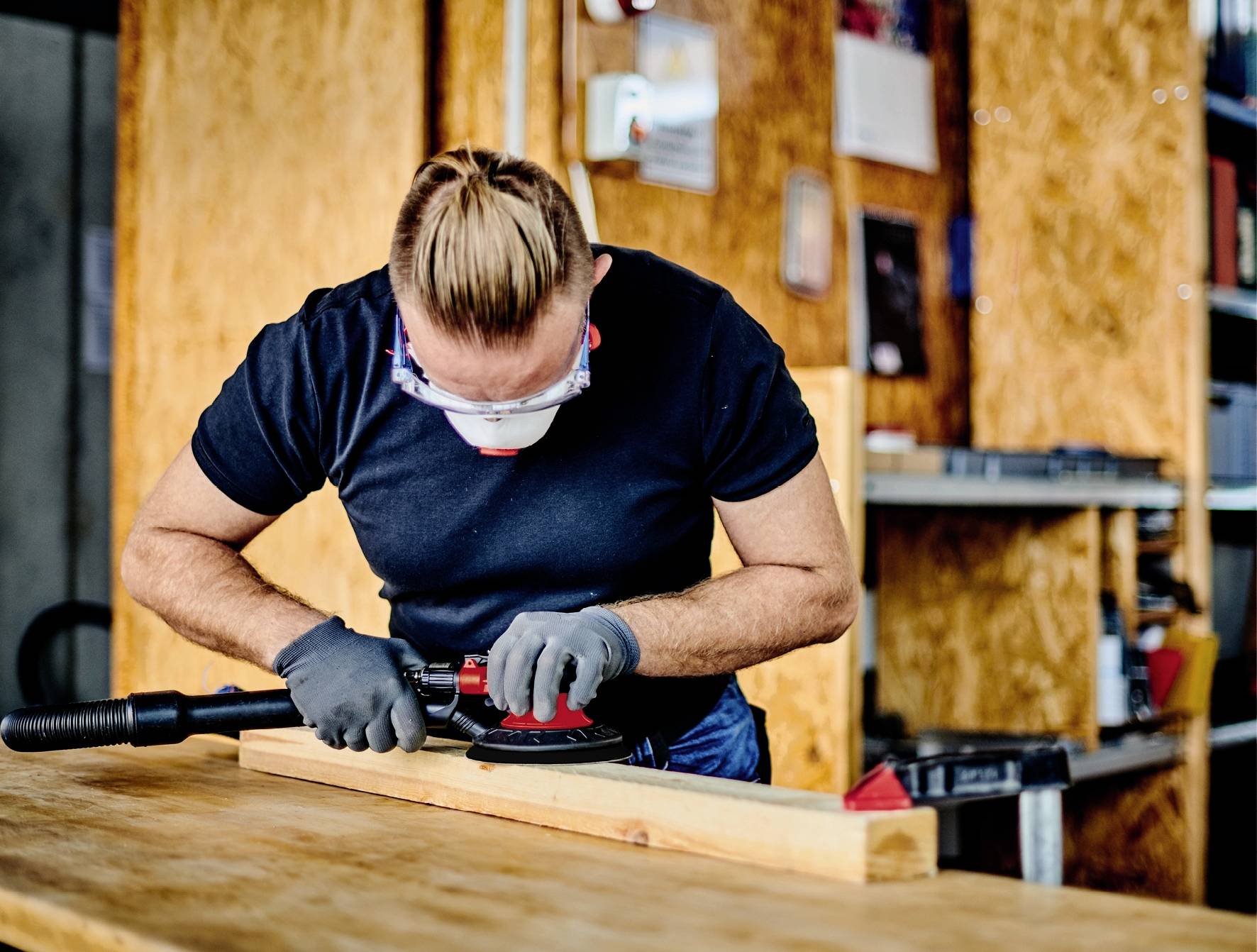 Eine Person schleift mit einer elektrischen Schleifmaschine ein Holzbrett in einer Werkstatt. Sie trägt Schutzbrille und Maske.