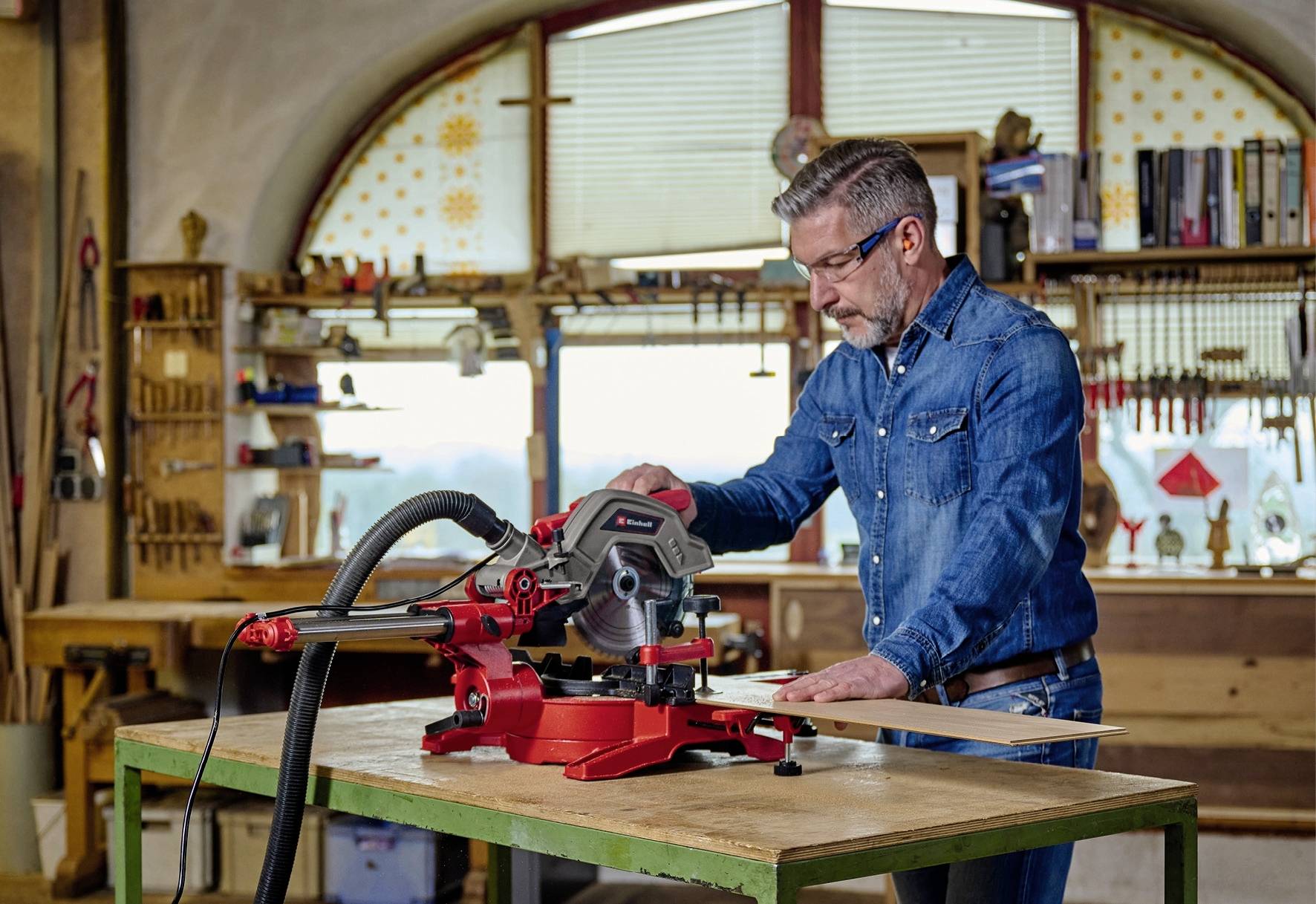 Ein Mann in Jeanskleidung bedient eine rote Tischkreissäge in einer Werkstatt mit Holzinventar und Werkzeugregalen im Hintergrund.